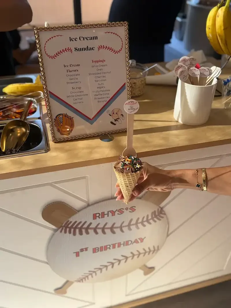 A person holding a chocolate ice cream cone with sprinkles, topped with a small white spoon with a message, in front of a sign listing ice cream flavors and toppings, and a baseball-themed cake with the name 'Rhys' and '1st Birthday' decoration, on a table at a birthday celebration.