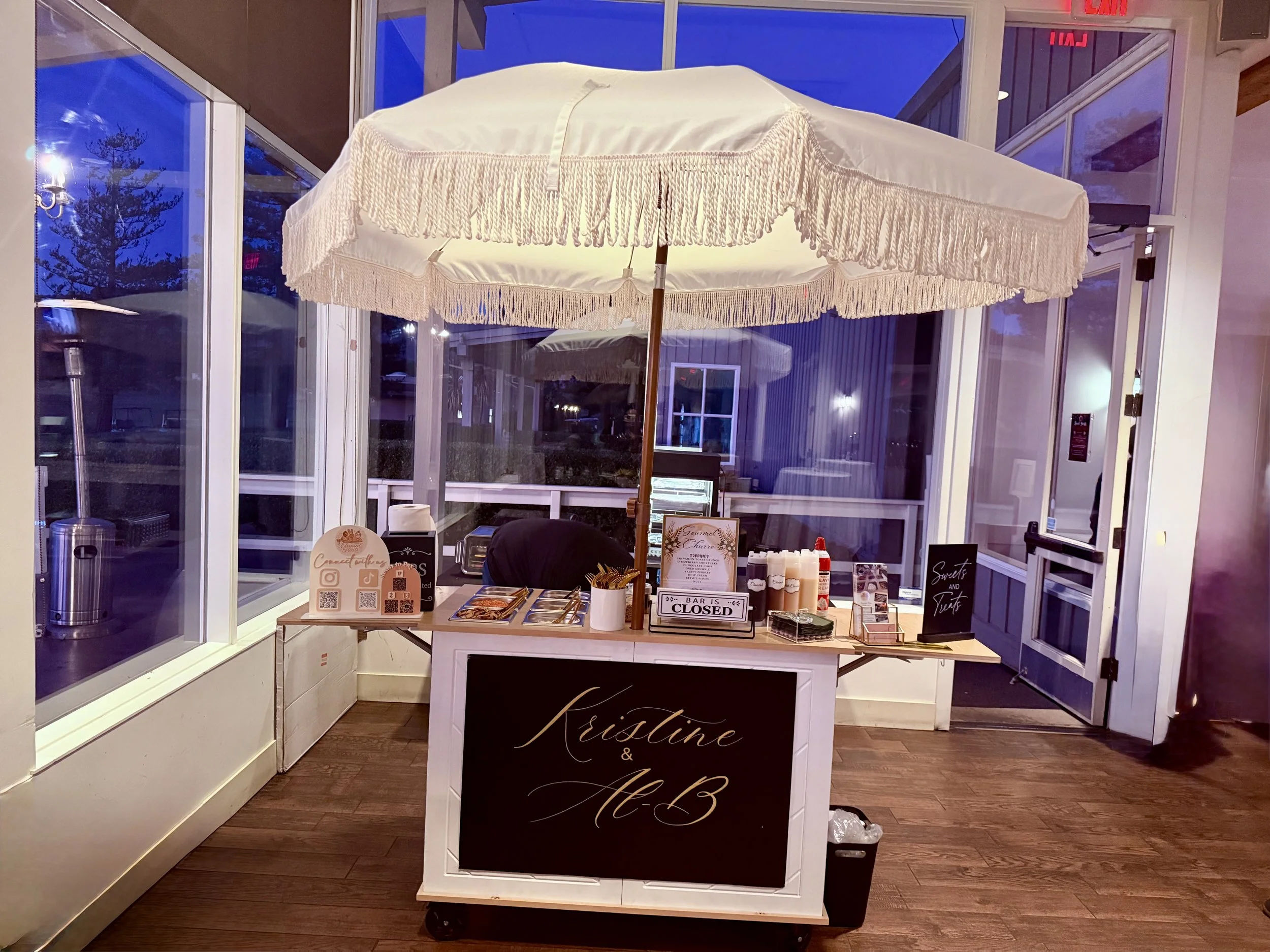 A small outdoor food or dessert stand with a white canopy and fringe, set up at night inside a glass-enclosed or glass-walled space. The stand has a black sign with white cursive writing reading 'Kristine & A.B.' and several bottles, flyers, and signs on top. A closed sign is visible on the front of the stand.