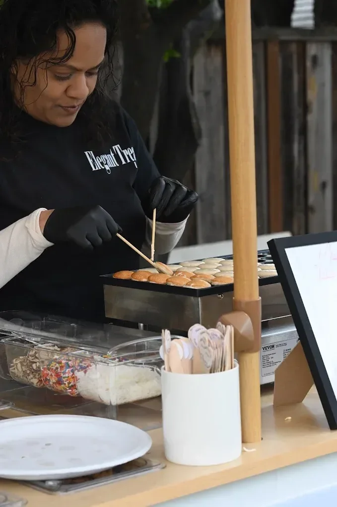 A woman and a child making and cooking round dough balls at a food stand. The woman is wearing a black shirt with white text and black gloves, using tongs to handle the dough. There are containers of sprinkles, sugar, and icing in front of them, and a tablet on the counter.