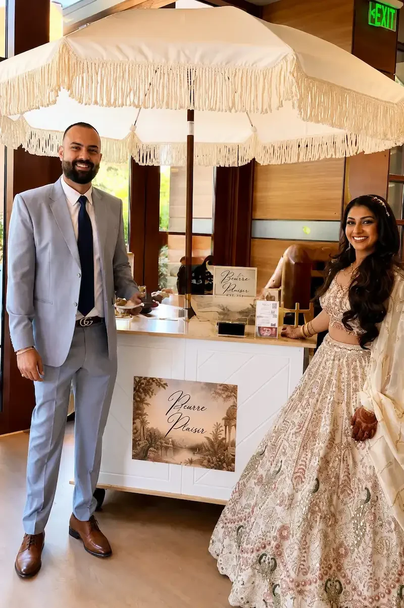 A man and woman in formal attire standing next to a dessert cart under a large cream-colored parasol inside a building. The man is wearing a light gray suit with a white shirt and dark tie, and the woman is wearing a traditional, ornate dress with intricate patterns. They are smiling at the camera. There is a sign on the cart that says "Beurre Plaisir" and an illuminated "EXIT" sign is visible in the background.