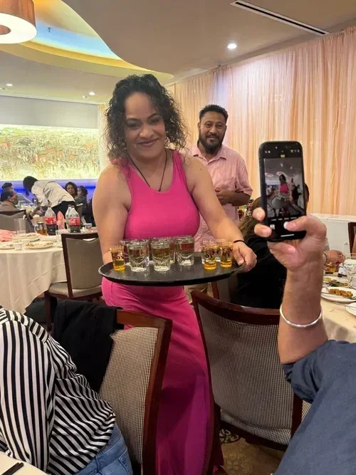 A woman in a pink dress serving drinks at a celebration or party in a restaurant, with people seated at tables and taking photos.