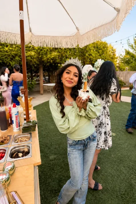 Young woman wearing a tiara and green top holding a dessert cup with a spoon at a backyard outdoor party.