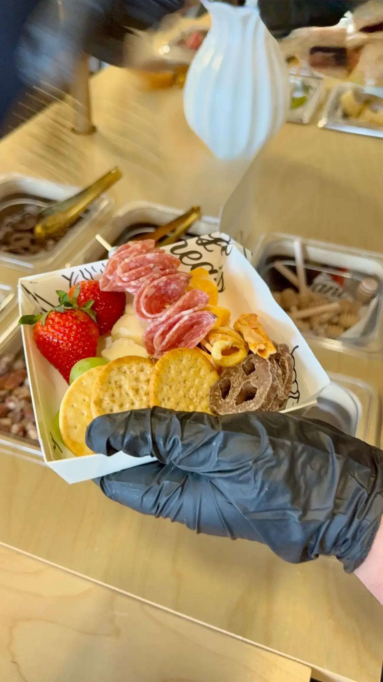 A person wearing a black glove holding a tray of assorted snacks and fruit, including strawberries, crackers, sliced meats, cheese, and chocolate chip cookies, with a background of condiment bottles and food containers.