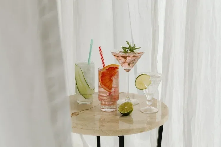 Six different colorful cocktails with garnishes on a round table in front of a white curtain.