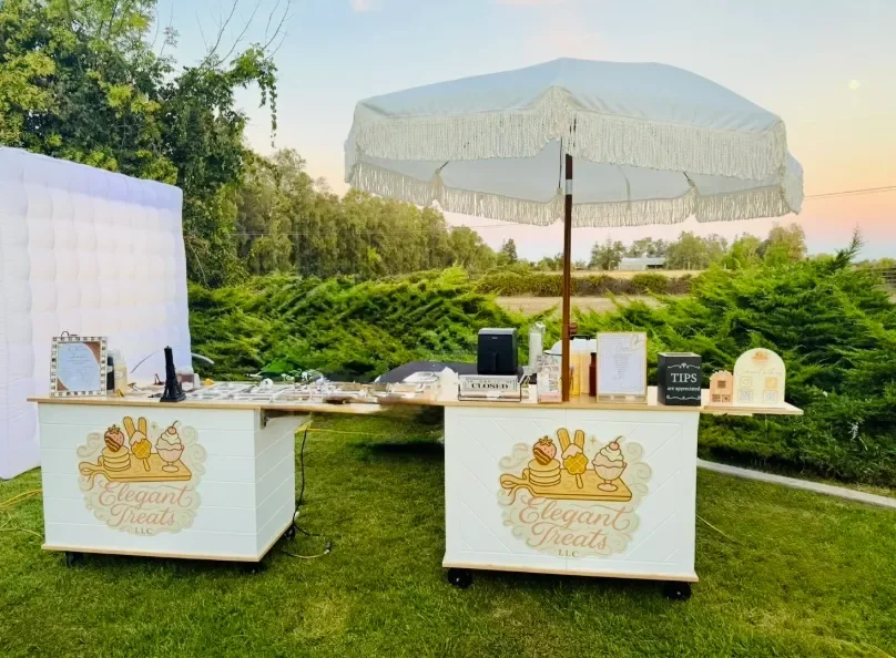 Outdoor dessert stand with a large white umbrella, set against a green landscape at sunset. The stand features a sign with a logo for 'Elegant Treats LLC' depicting various desserts.