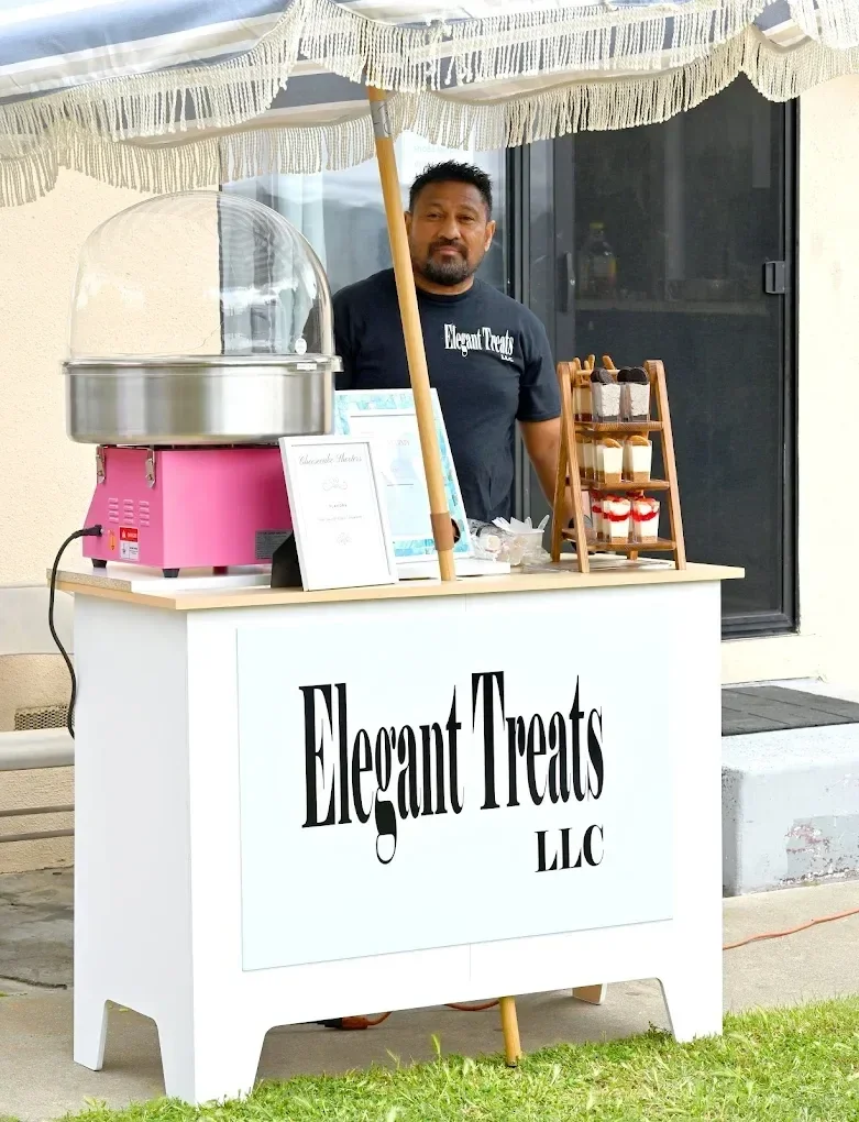 A man standing behind a dessert stand with a sign that reads 'Elegant Treats LLC'. The stand has a cotton candy machine, a small menu, and containers of treats, all shaded by a large fringed umbrella.