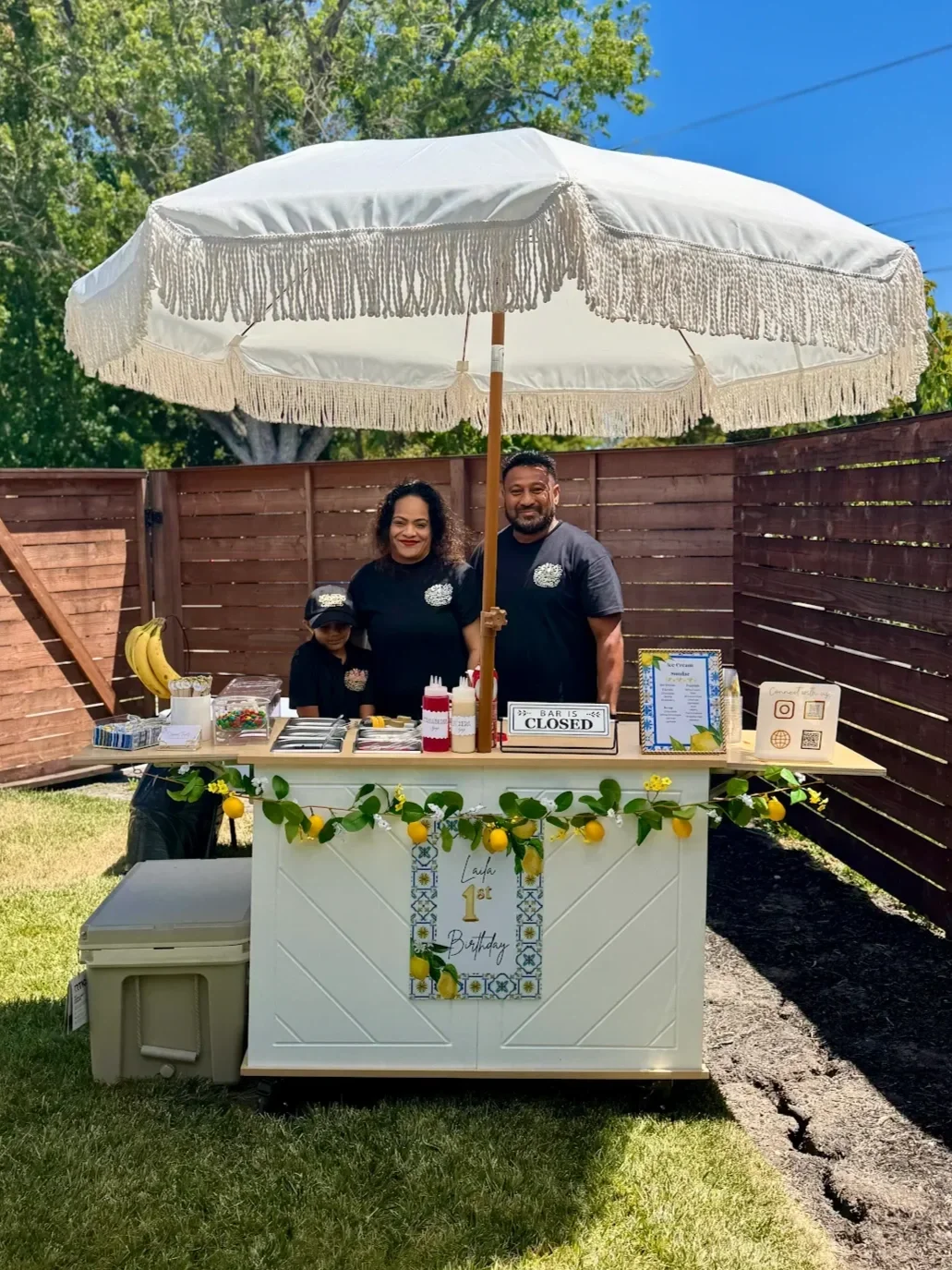 Family standing behind a lemon-themed birthday party food cart outdoors under a large white parasol, with a sign indicating the bar is closed.