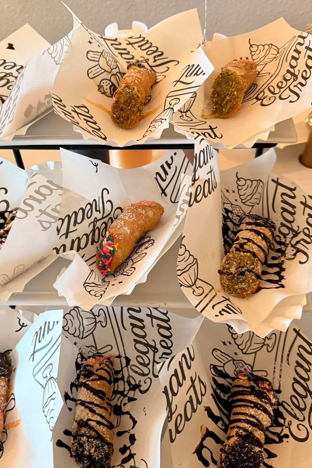 Assorted cannoli, some decorated with sprinkles and chocolate drizzle, resting on printed paper liners.