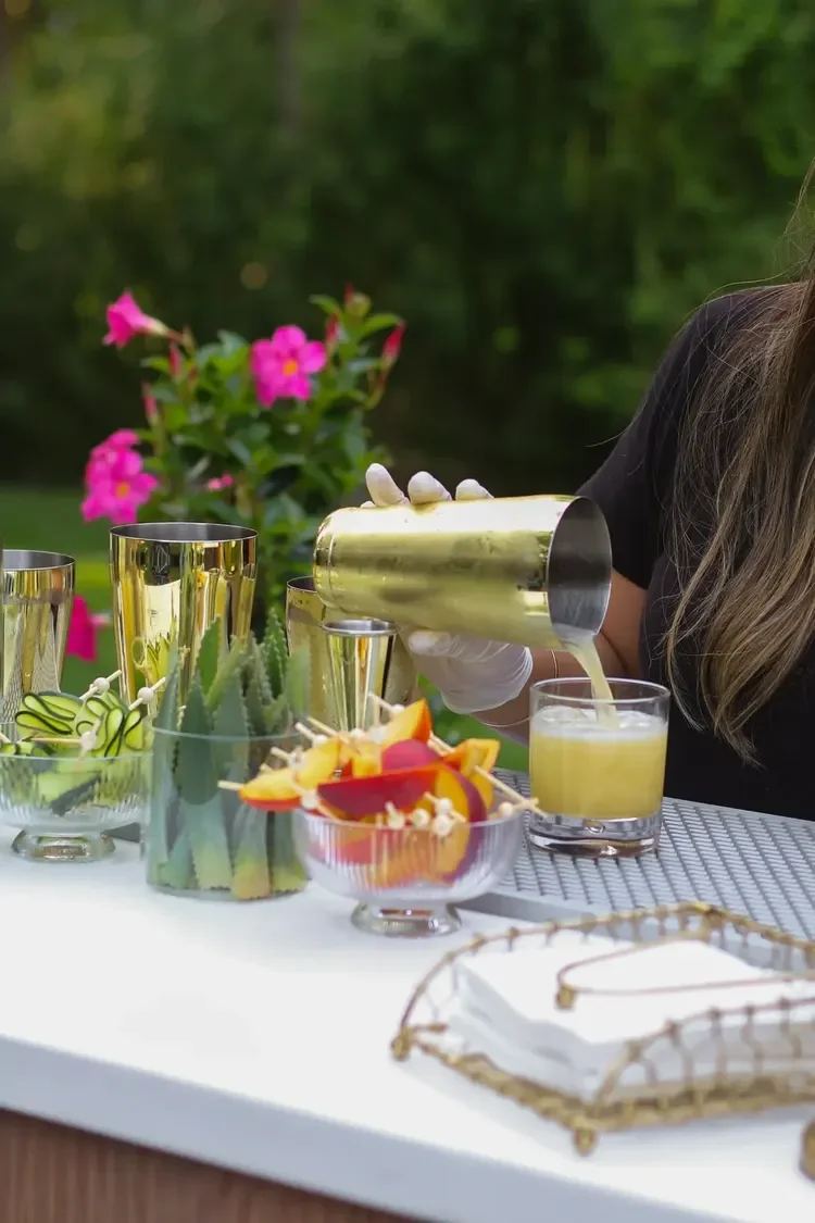 Person pouring a yellow beverage into a glass on an outdoor table with various bowls of fruit and decorative items