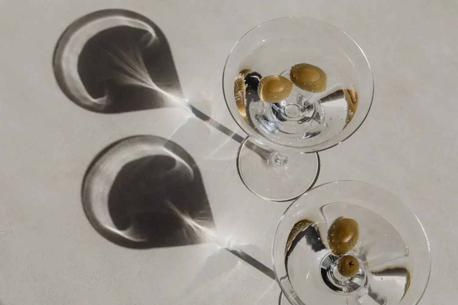 Two empty cocktail glasses with green olives inside, casting shadows on a light surface.