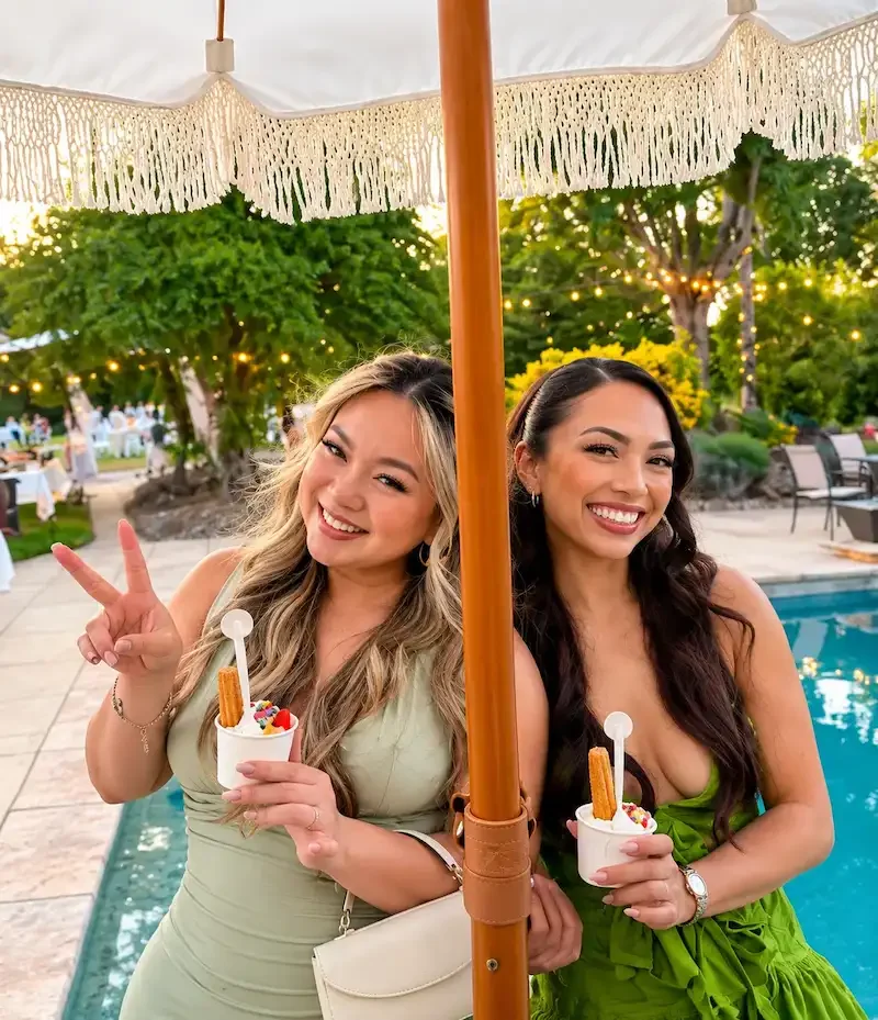 Two women smiling and holding cups of ice cream or frozen yogurt, standing by a poolside beneath a cream-colored parasol, during dusk with string lights and greenery in the background.