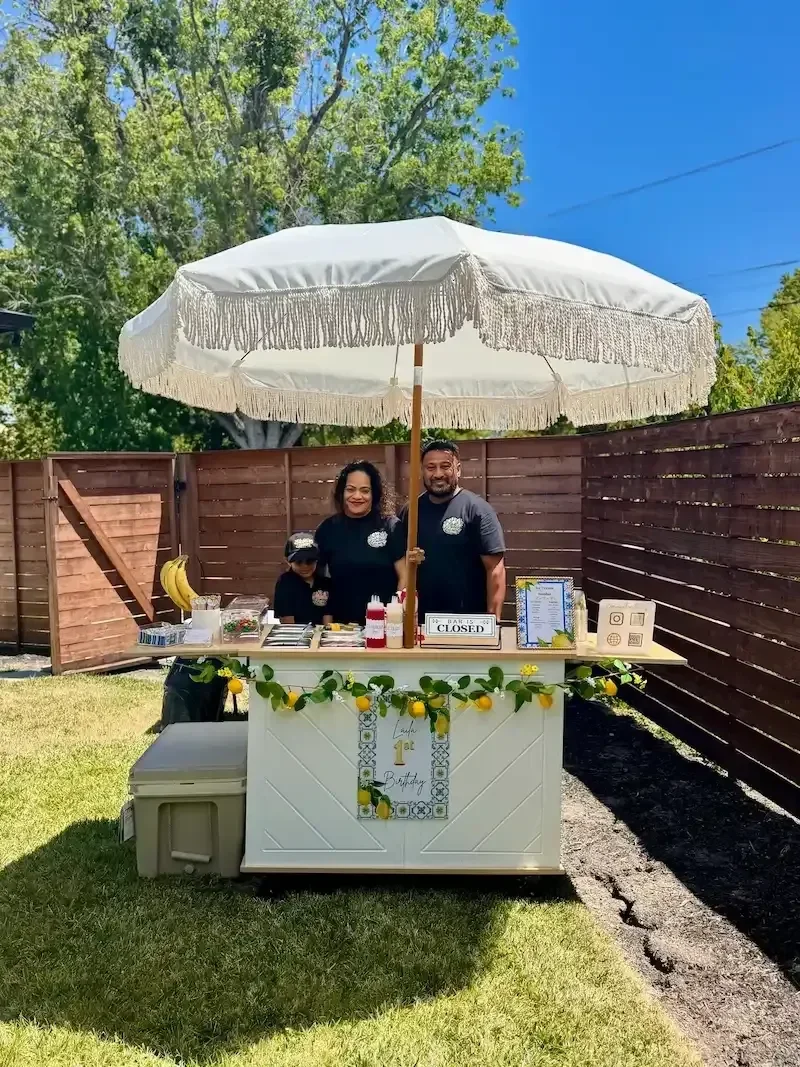 A family of three standing behind a white lemonade stand under a large white umbrella in a backyard with a wooden fence and green trees, celebrating a birthday with lemons and bananas on the stand.