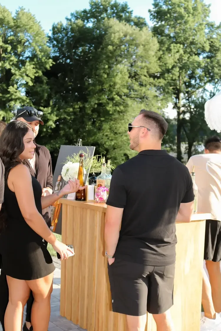 People at an outdoor bar with trees in the background, drinks on the bar, and a woman in a black dress talking to a man in sunglasses and a black shirt.
