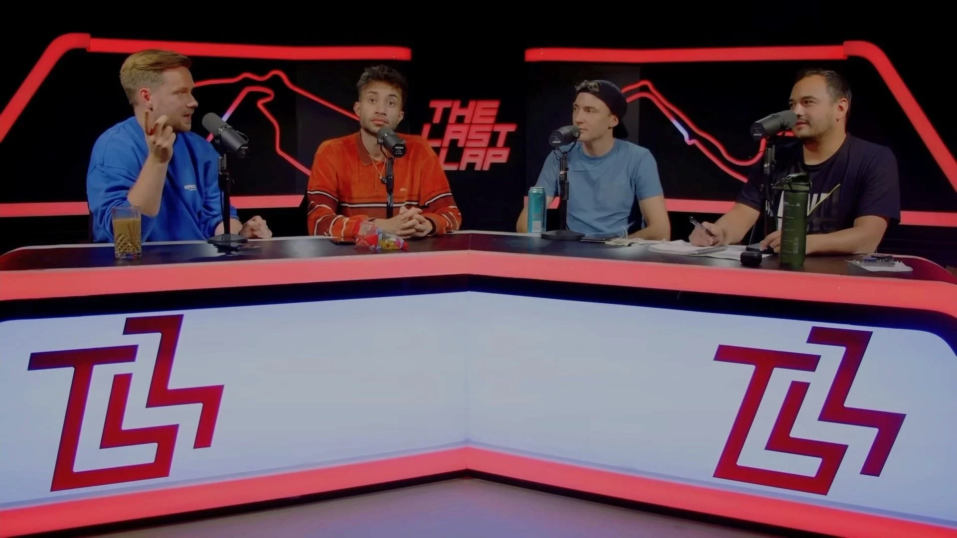 Four men sitting at a podcast table with microphones, drinks, and snacks, in front of a backdrop with a neon red line graph and the text "The Last Lap."