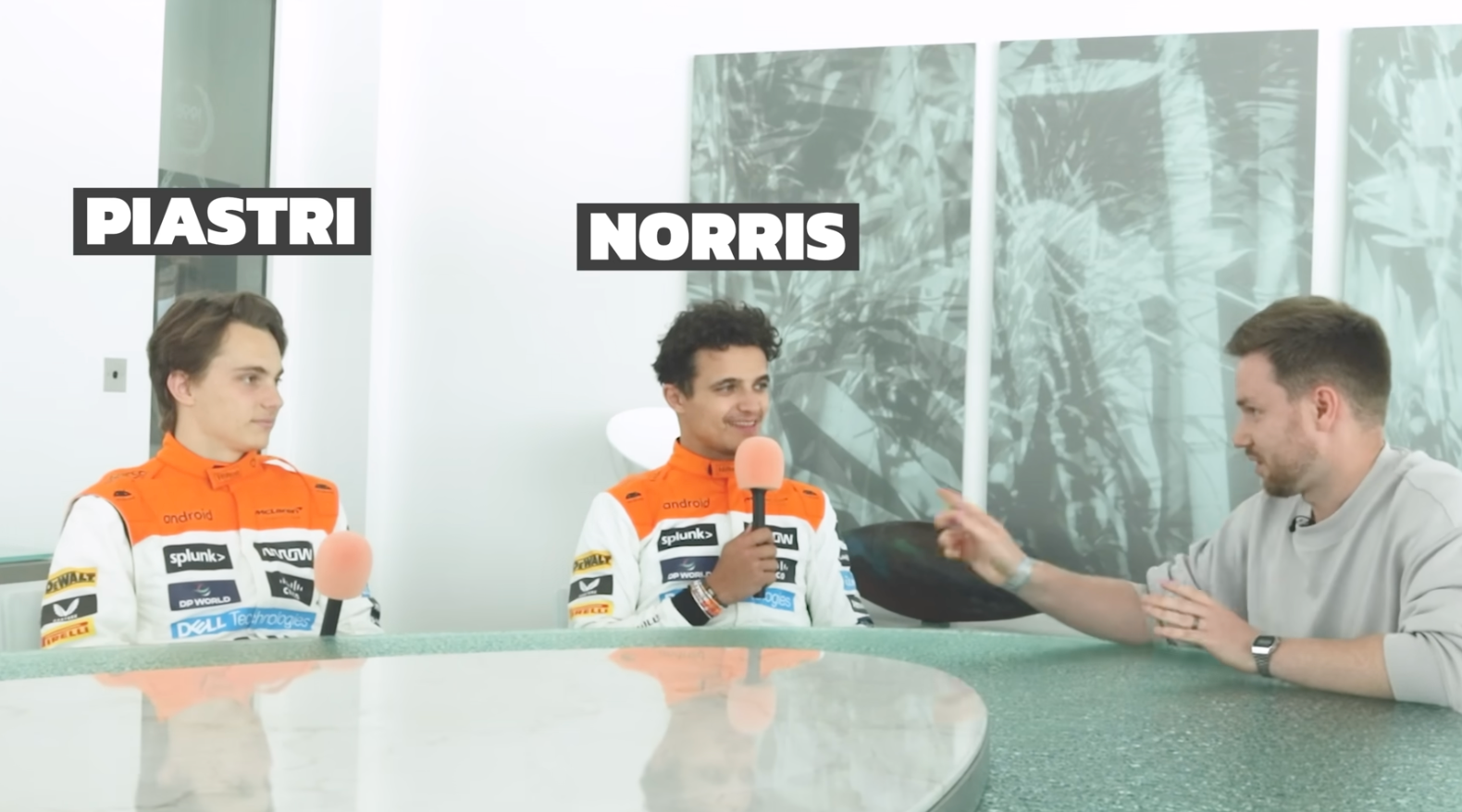 Three race car drivers in orange and white racing suits sitting at a green table in an interview setting, with a man on the right pointing at one of the drivers.