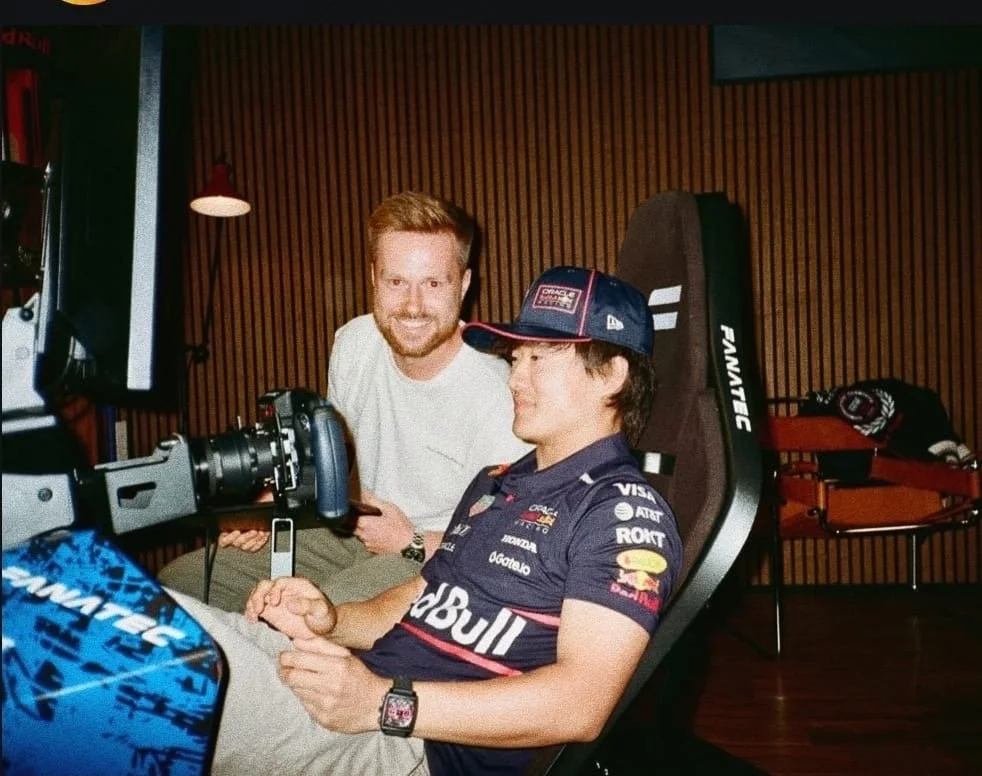A man in a Red Bull Racing Team shirt sitting in a gaming chair, looking at a camera with a smile, while another man with a white shirt stands beside him, smiling. The background has wooden paneling and a small table with merchandise or gear.