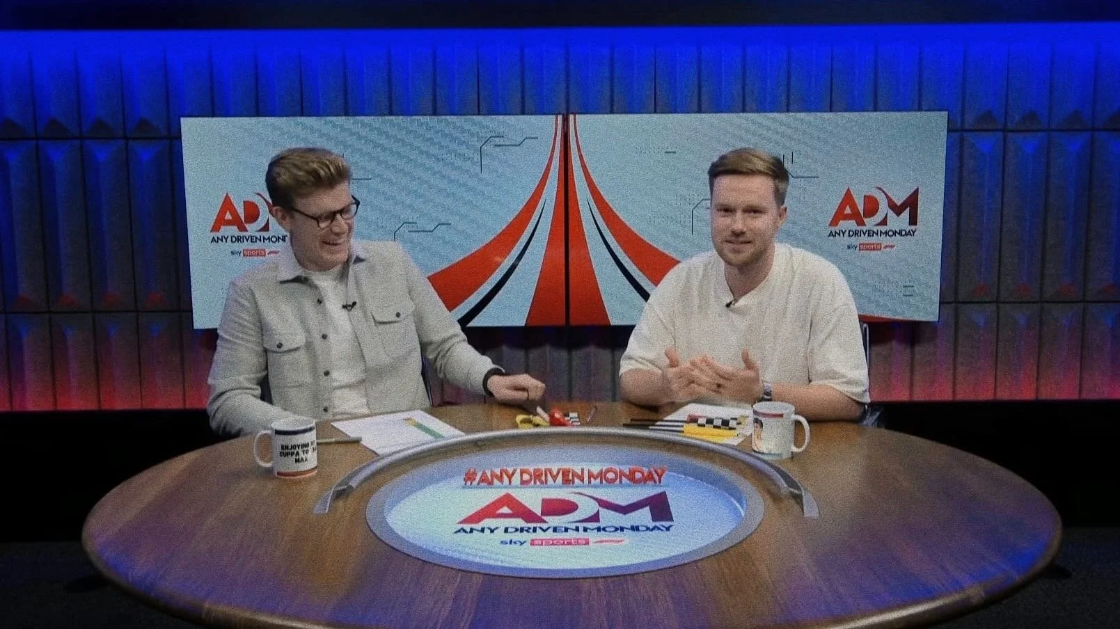 Two men sit at a round table in a TV studio with a backdrop displaying the logo for 'AD' and the hashtag '#ANYDRIVENMONDAY.' The man on the left wears glasses and a light gray jacket, while the man on the right wears a white shirt. There are mugs, papers, and small objects on the table.