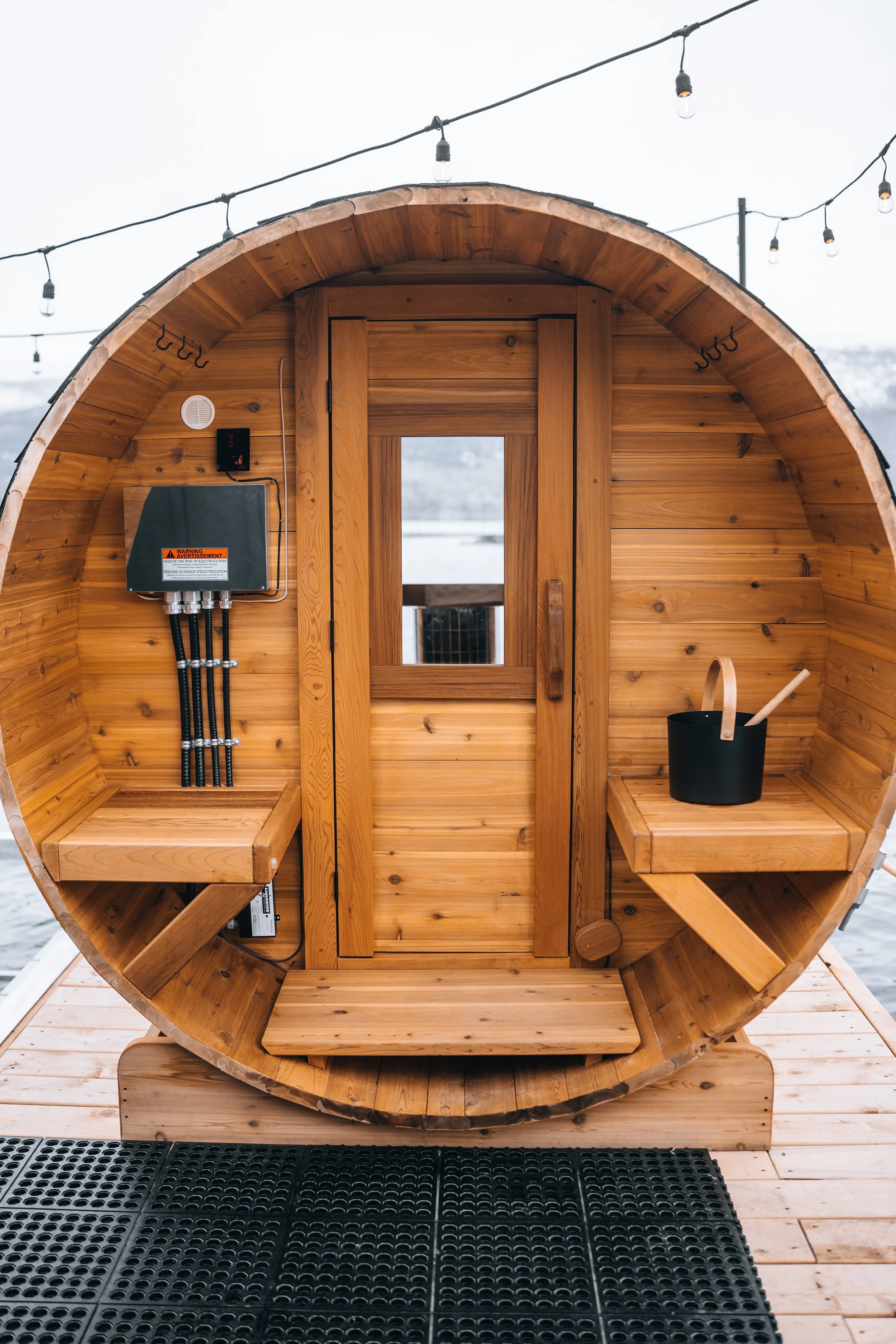 A small, circular wooden sauna with string lights overhead and a wooden deck outside.