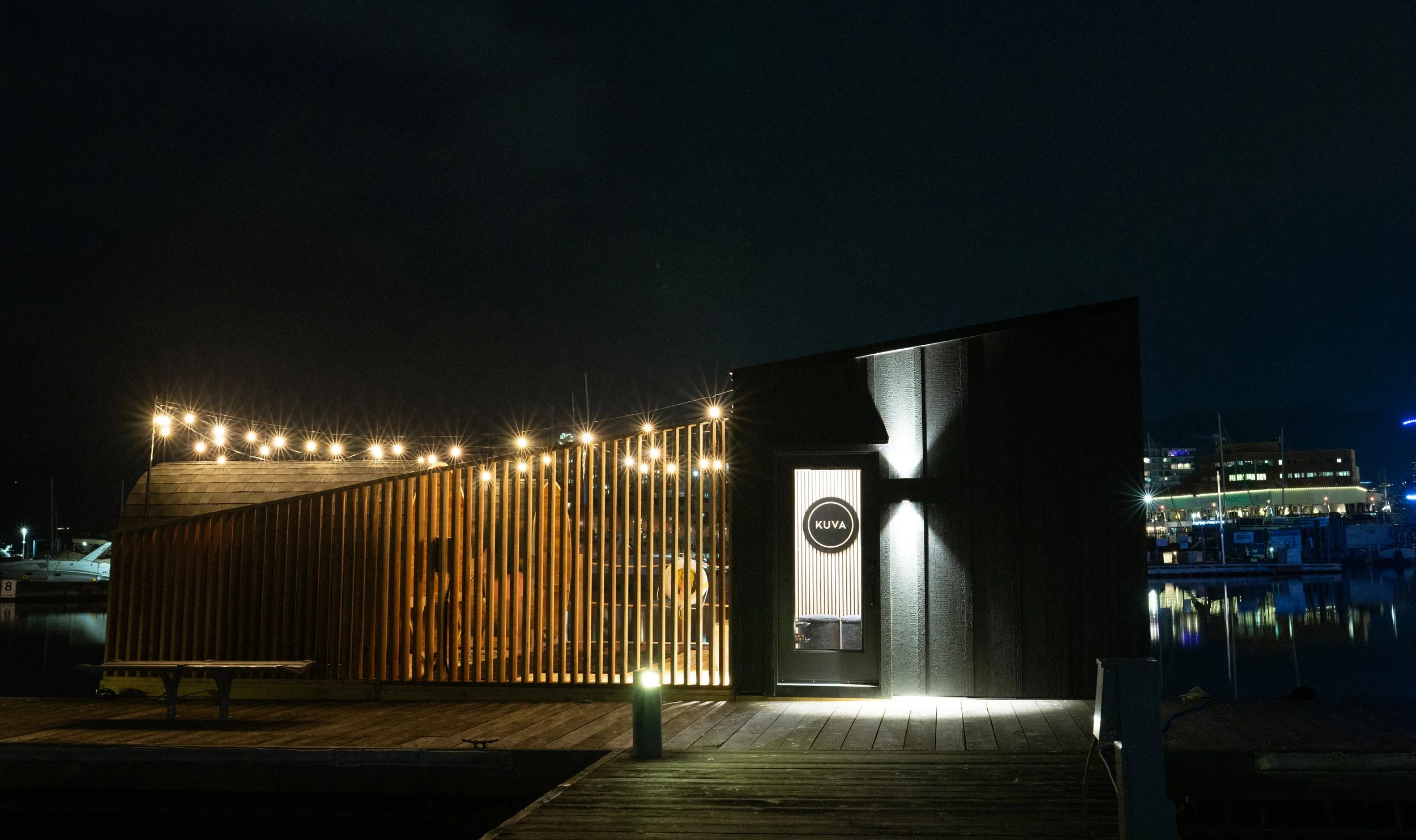 Night view of a modern building with a curved wooden facade and illuminated sign that reads 'KUVA', next to a door, with boats docked in the background and outdoor string lights.