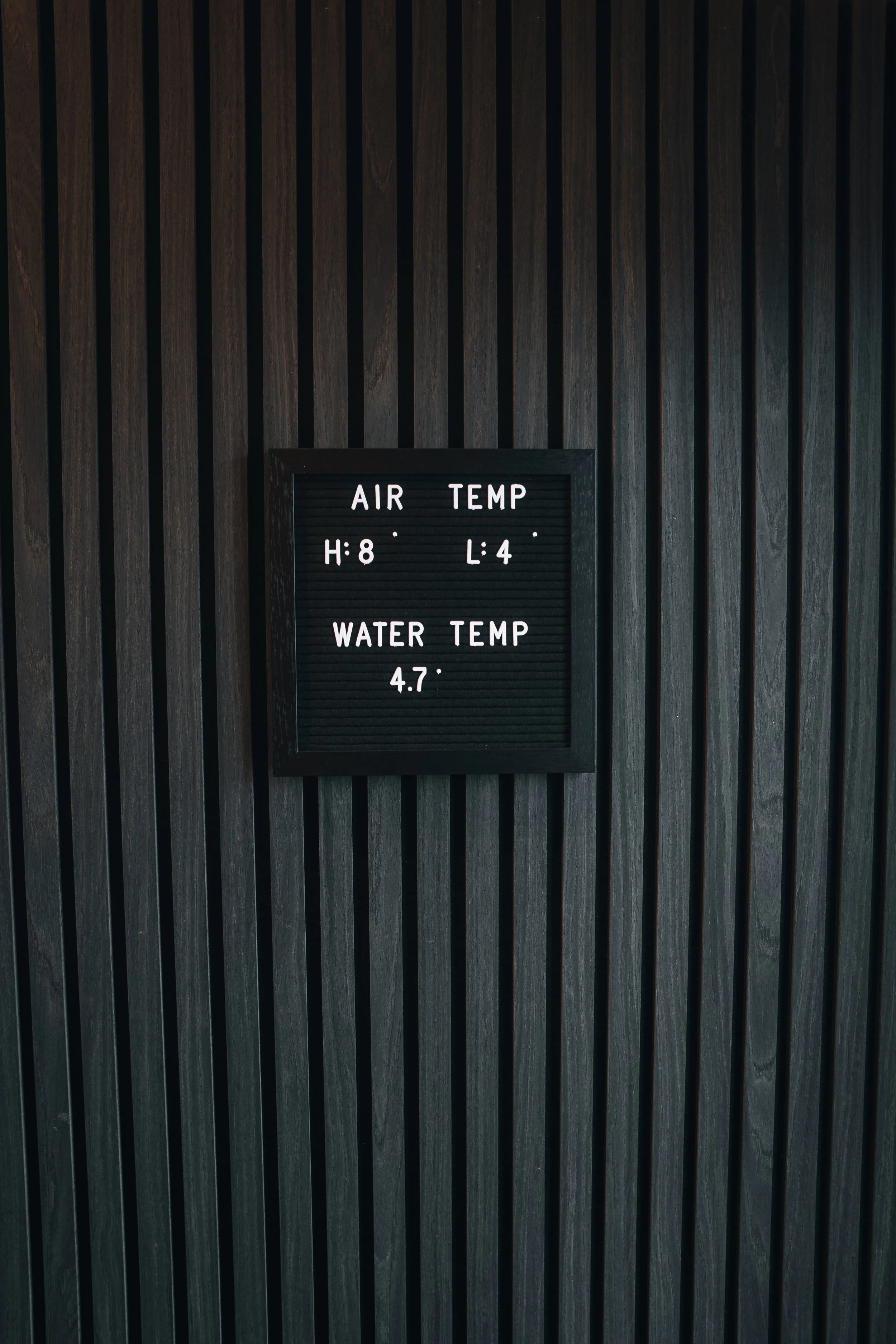 Black display board with white letters showing air and water temperatures on a dark wooden wall.