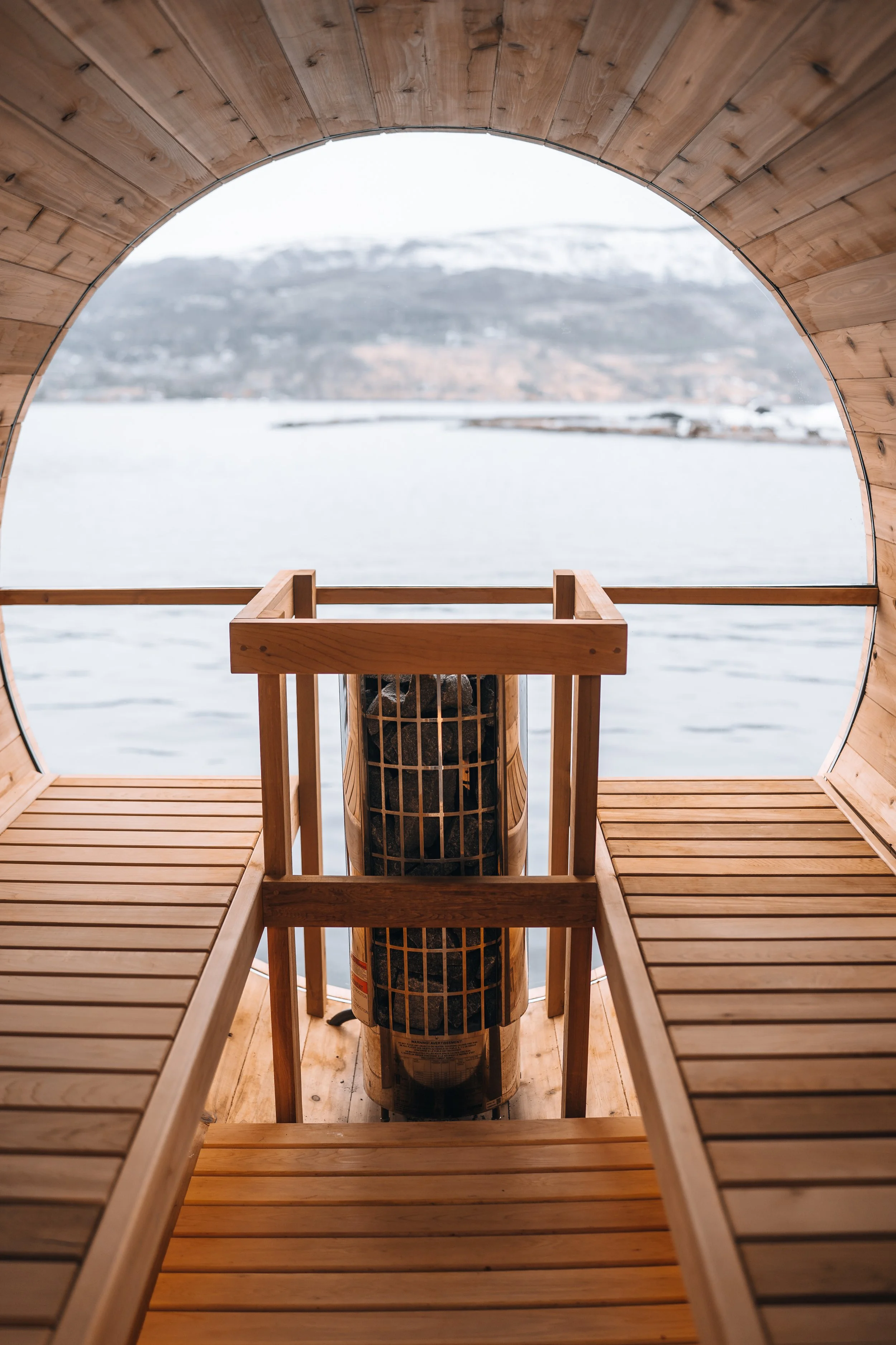View through a circular window of a wooden sauna overlooking a body of water and distant hills or mountains.