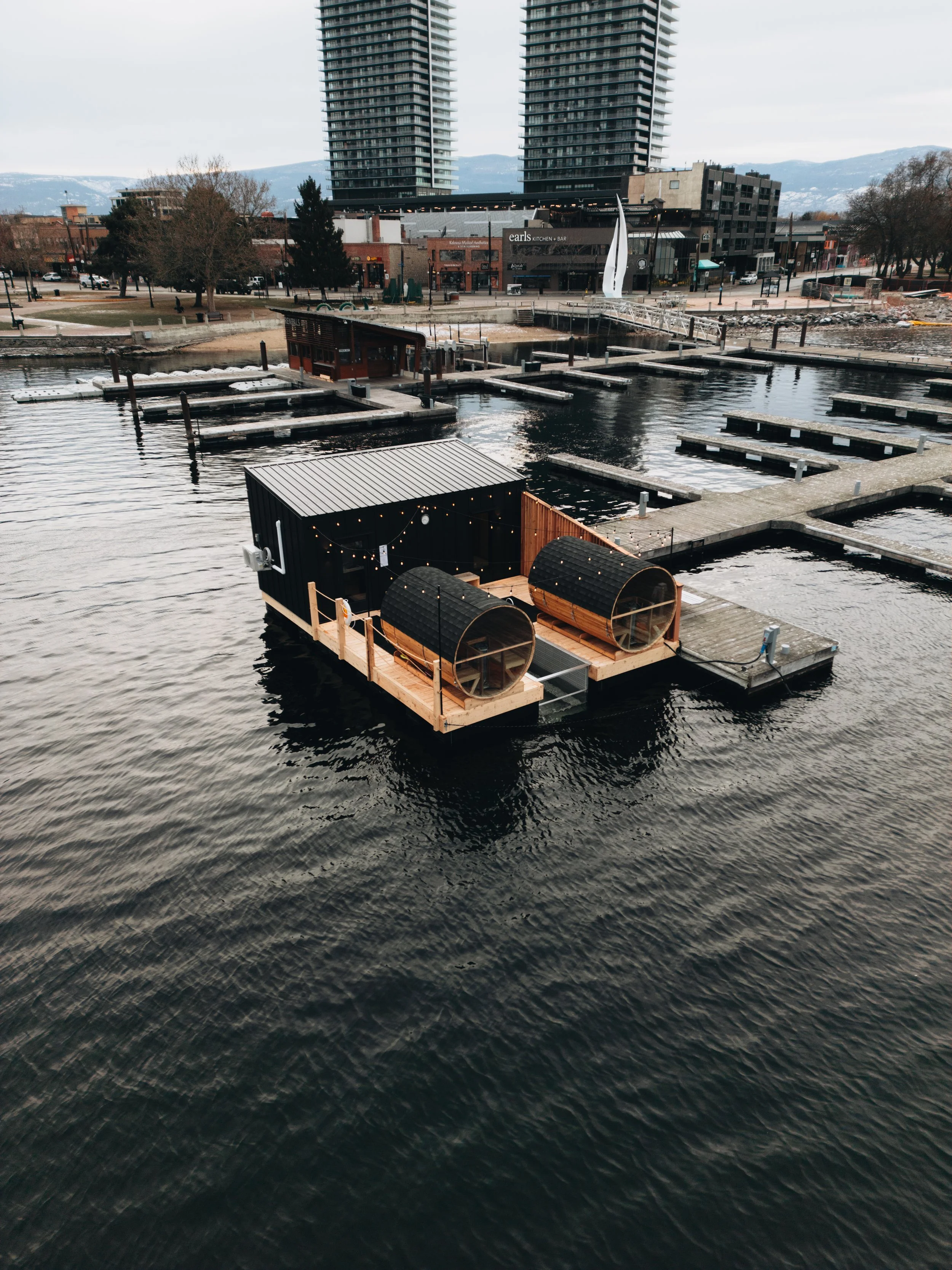 A floating dock with two enclosed saunas, decorated with string lights, on a marina with empty boat slips. In the background are urban buildings and two tall residential towers, with mountains visible in the distance.