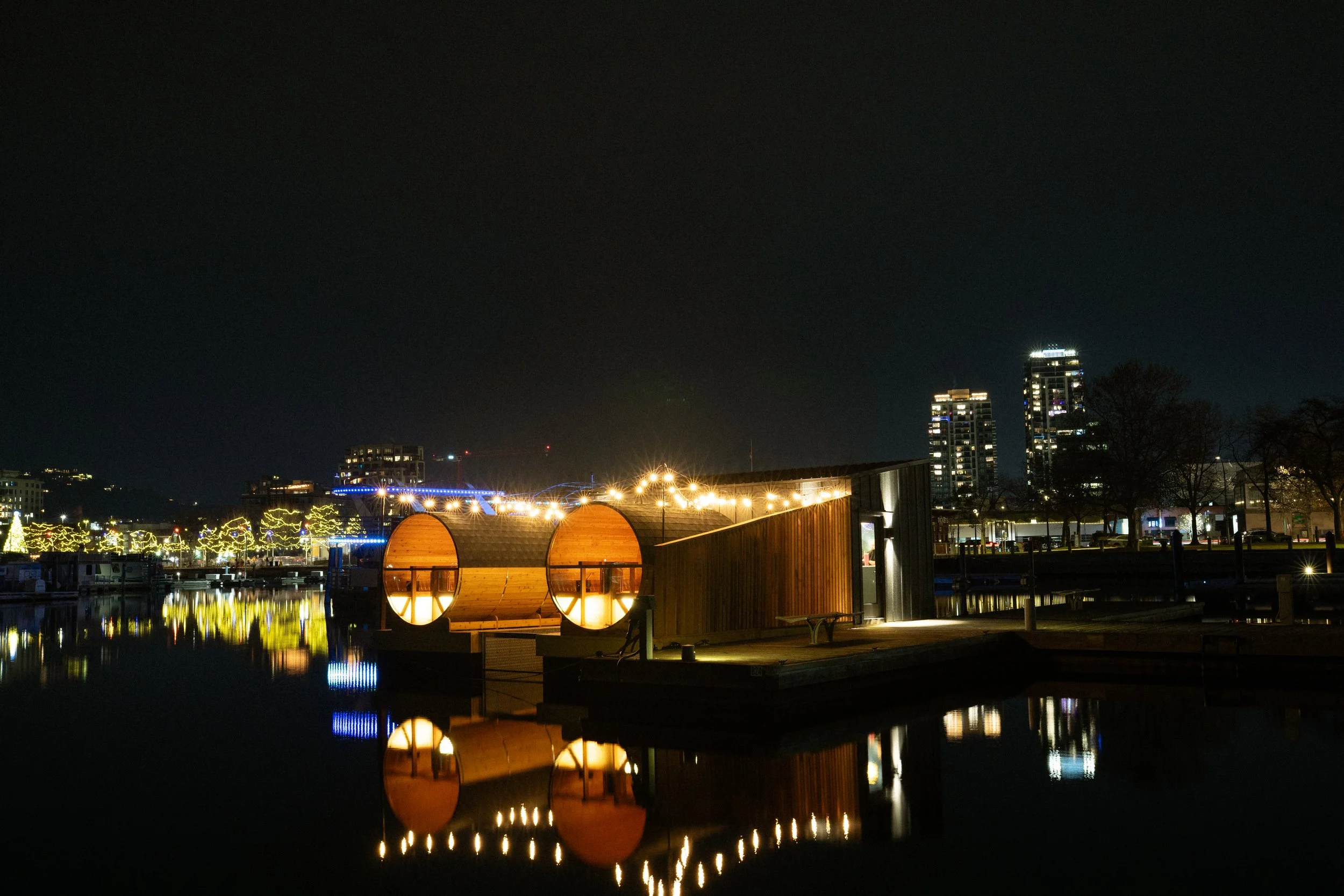 Nighttime view of a modern waterfront pavilion with circular design, illuminated by string lights, reflected in the calm water, with city buildings and trees in the background.