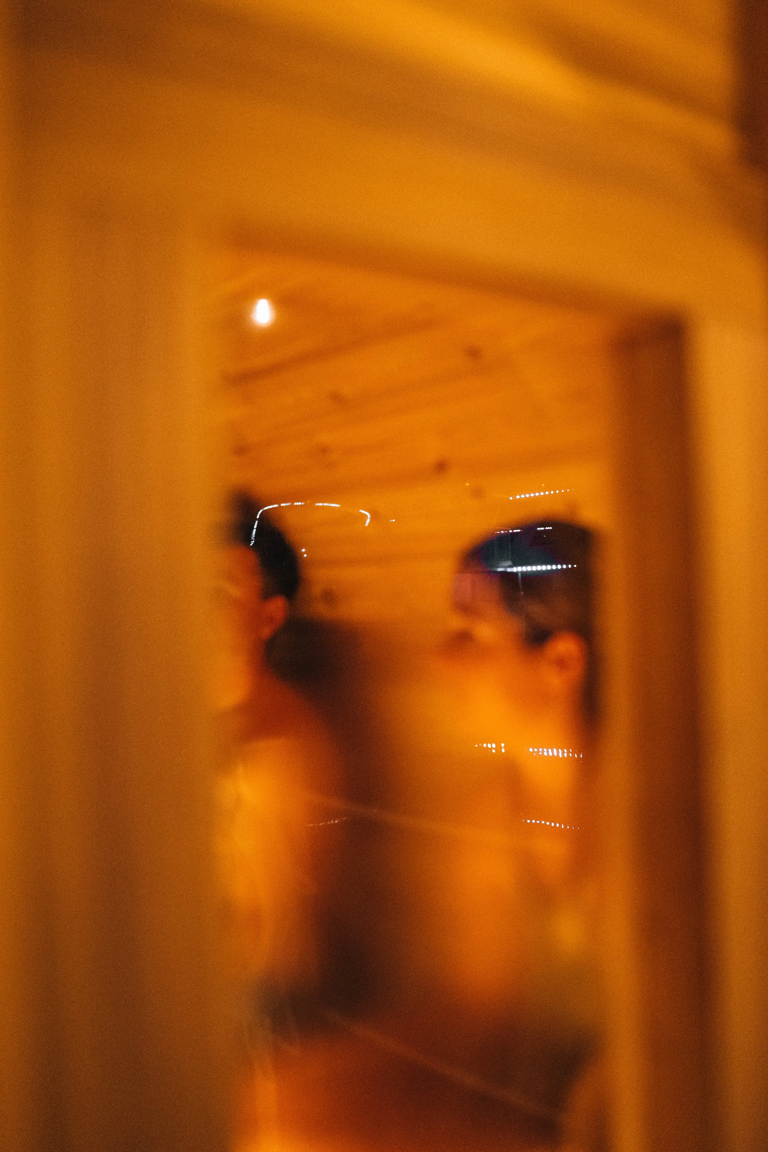 Reflection of a person's face in a mirror with wooden wall background, seen through a slightly blurred vertical frame.