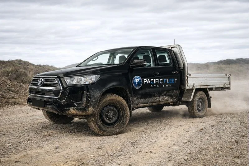 Black Toyota pickup truck with Pacific Fleet Systems logo, driving on a dirt road in a rural area, kicking up dust.