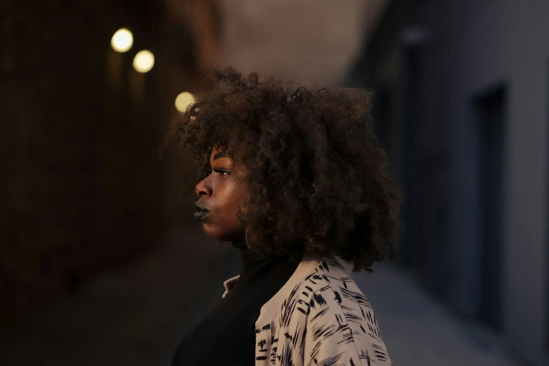 Side profile of a woman with curly black hair standing in an urban alleyway at night, illuminated by soft streetlights.