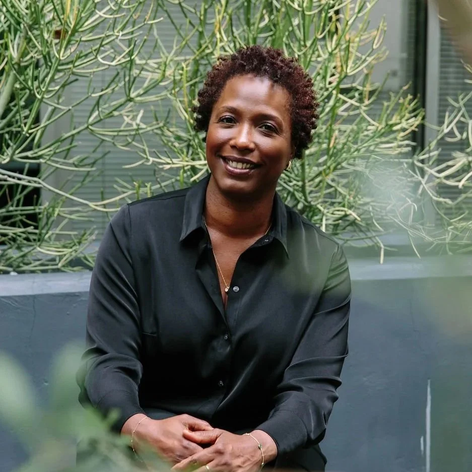 A woman with short curly hair smiling, wearing a black shirt, sitting outdoors in front of green plants.