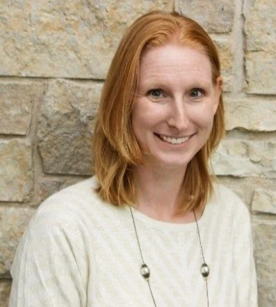 Close-up of a man with glasses and a nose ring, wearing a dark green sweater, looking to the side with a thoughtful expression, in a bright room with a plant in the background.