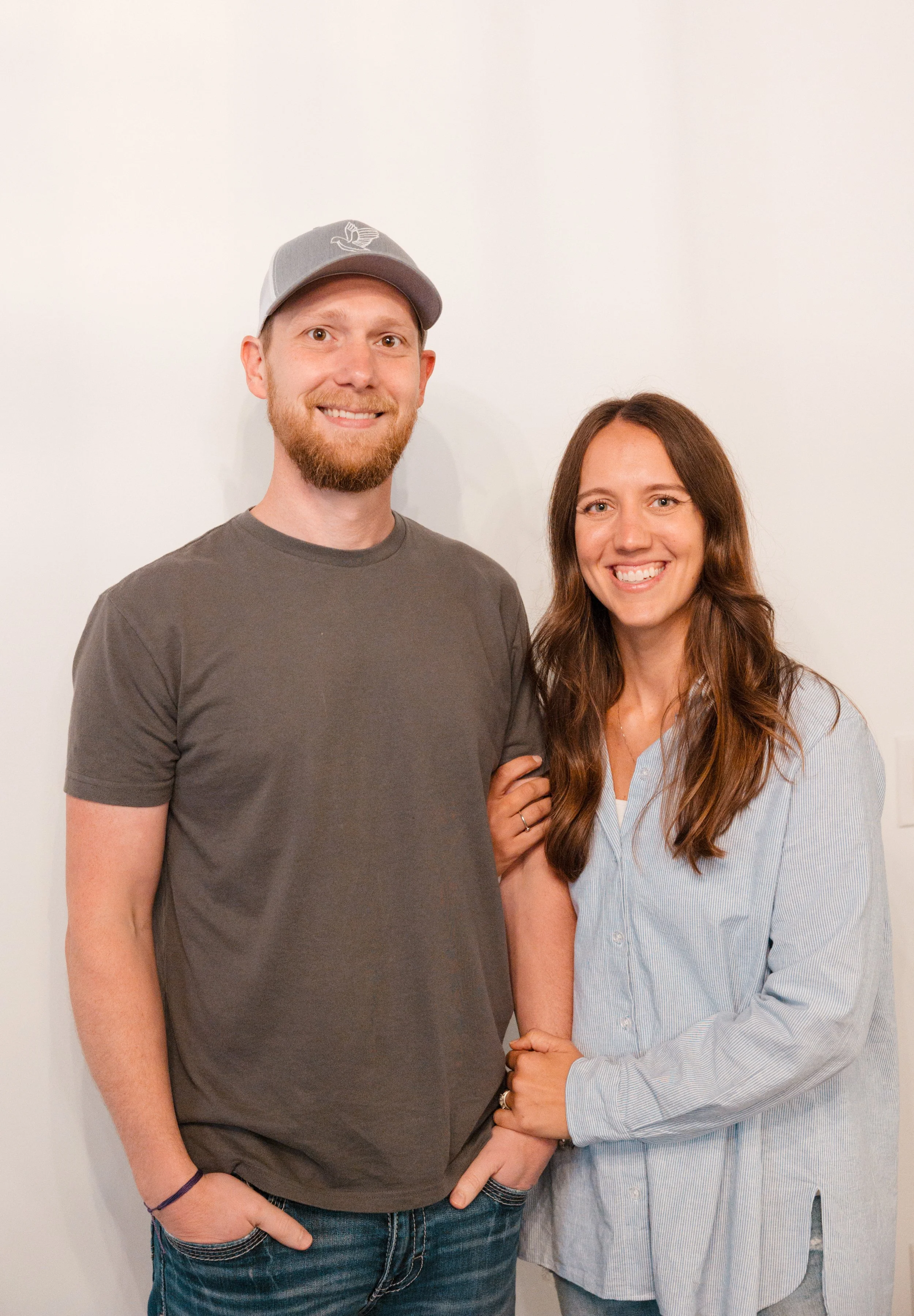 A smiling man in a grey cap and t-shirt standing next to a smiling woman in a light blue shirt, against a plain white wall.