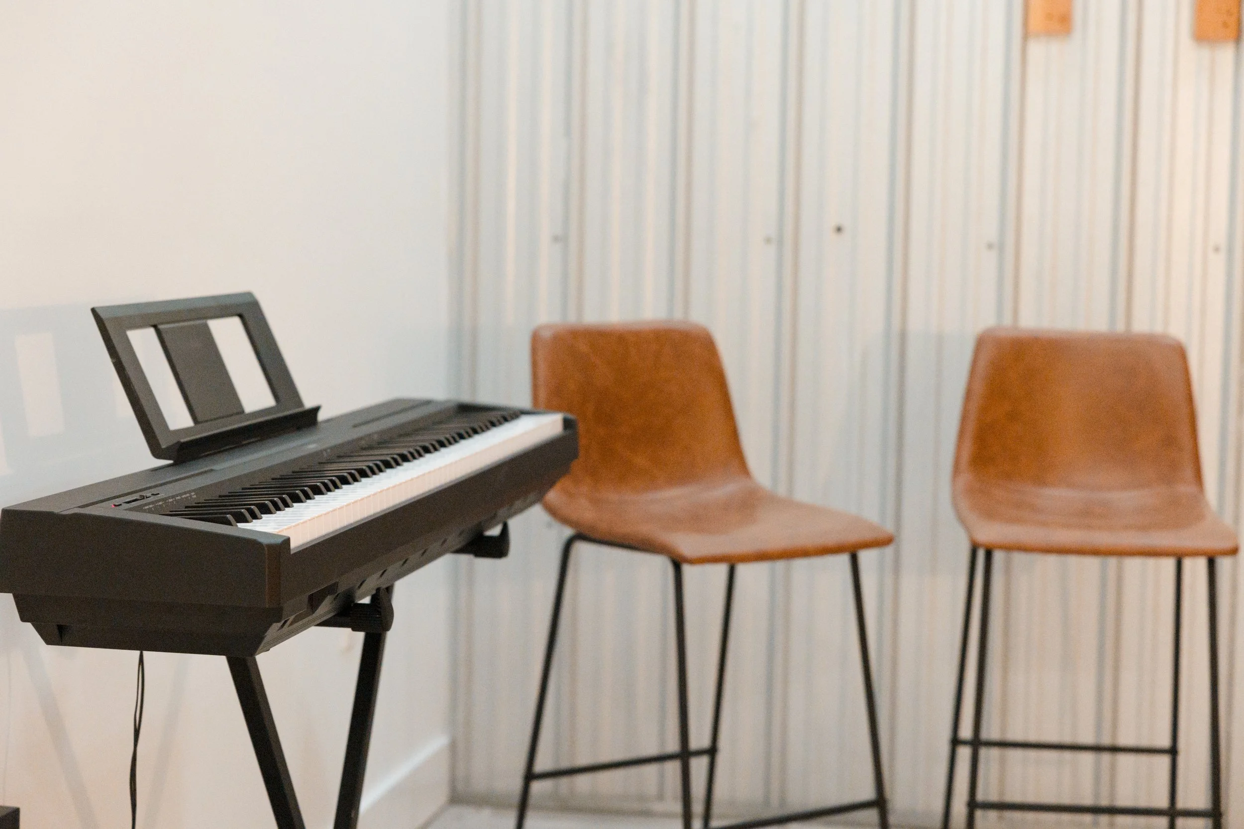 A digital keyboard on a stand next to two empty wooden chairs against a striped wallpaper background.  Music classes songwriting classes worship leading classes. 