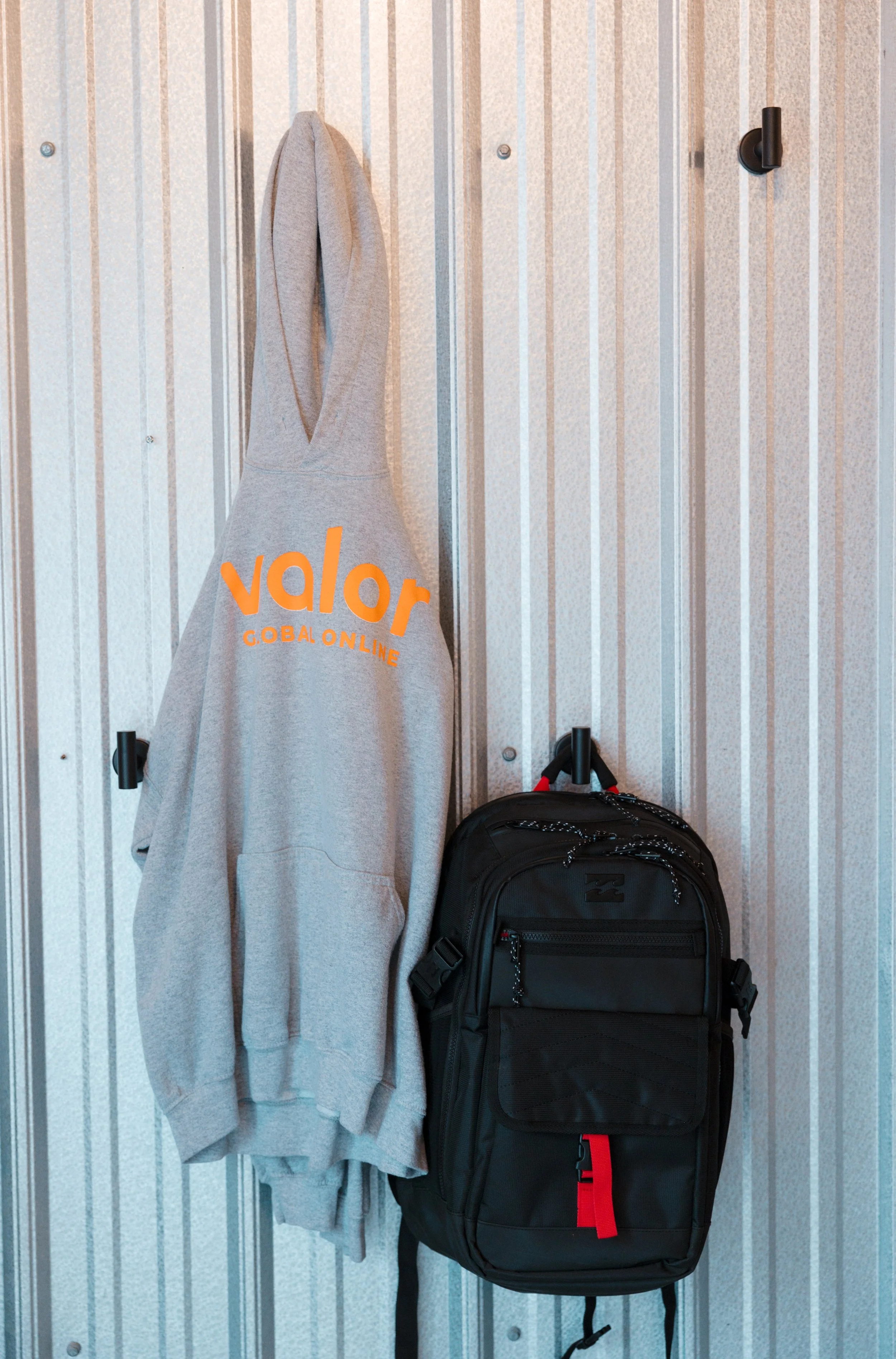 Gray hoodie with 'valor' logo hanging on a hook next to a black backpack on a striped metal wall.