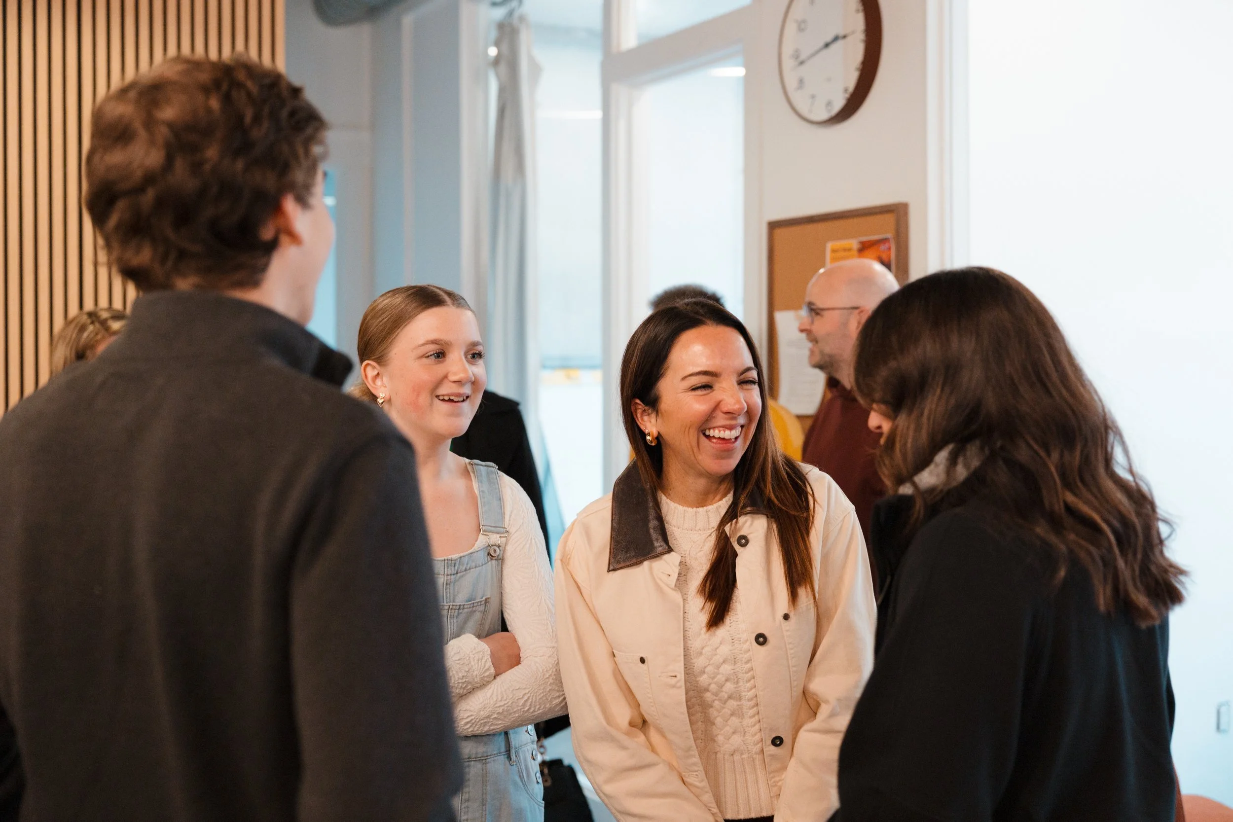 Group of five people talking and smiling in a room with a wooden wall, a window, and a clock.