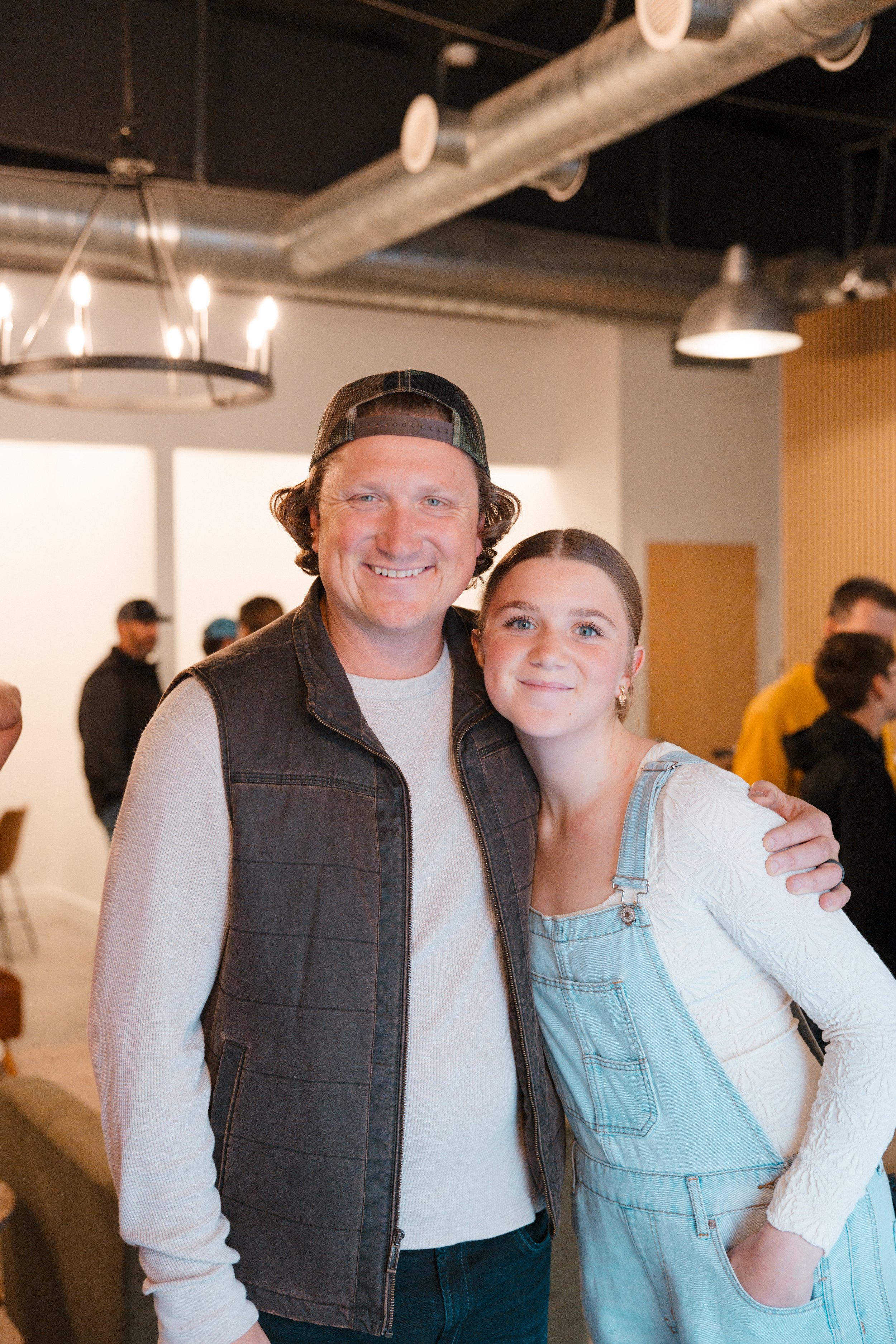 A smiling man in a black vest and baseball cap stands next to a girl in a white shirt and light blue overalls, both with arms around each other, inside a modern, industrial-style indoor space with warm lighting and other people in the background.