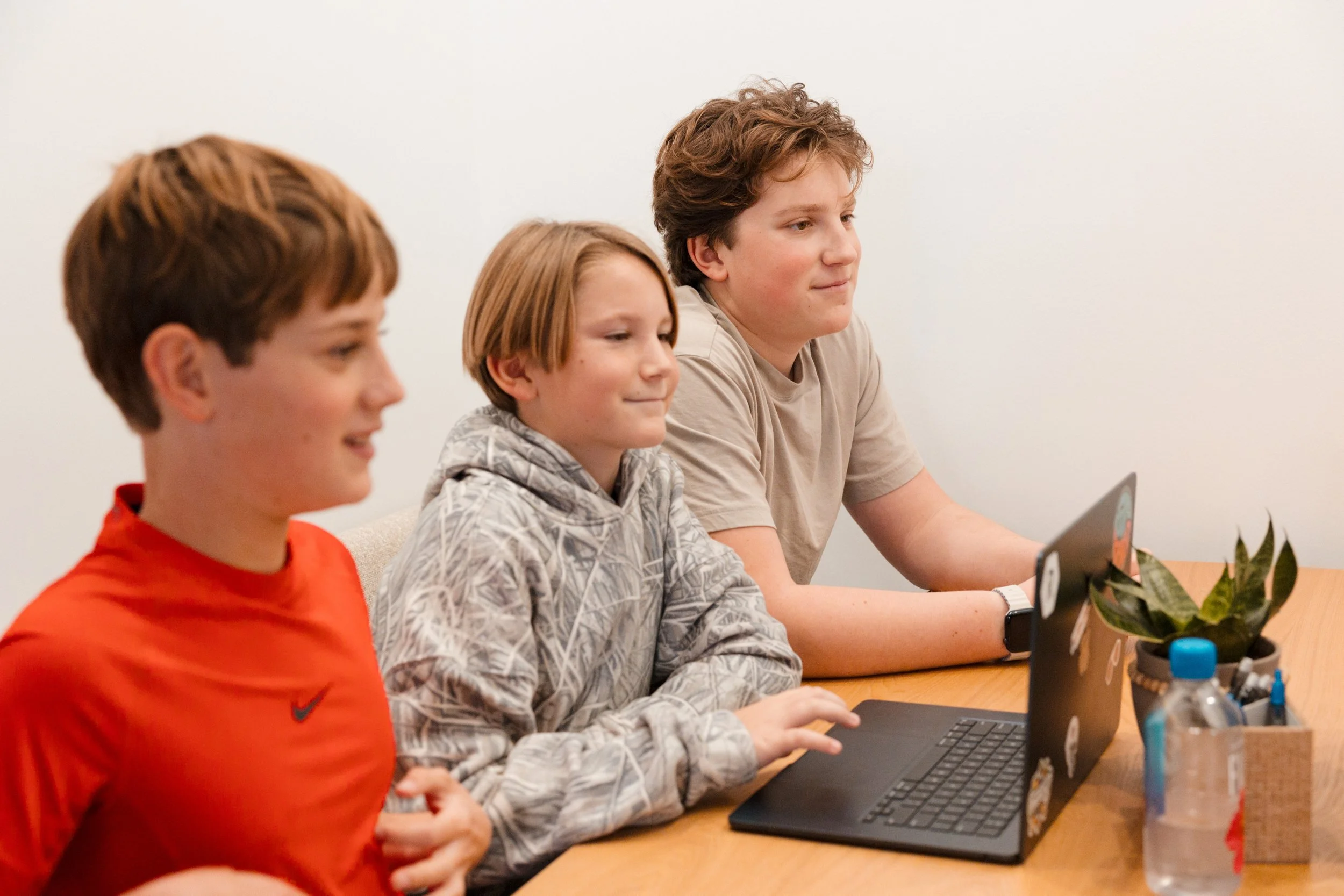 Three boys sitting at a wooden table in front of a laptop, with a potted plant and water bottles nearby.