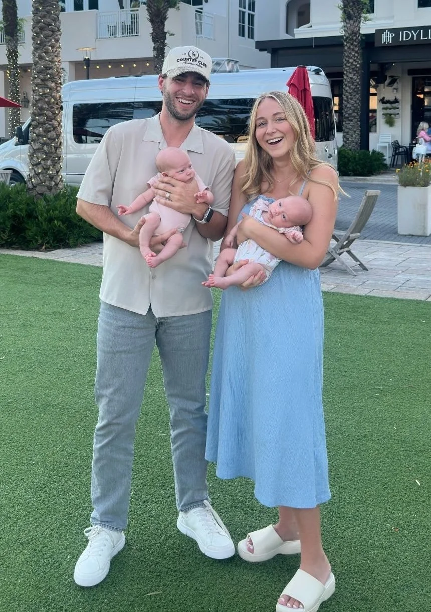 A man and woman smiling outdoors, each holding a baby, with palm trees, a building, and a van in the background.