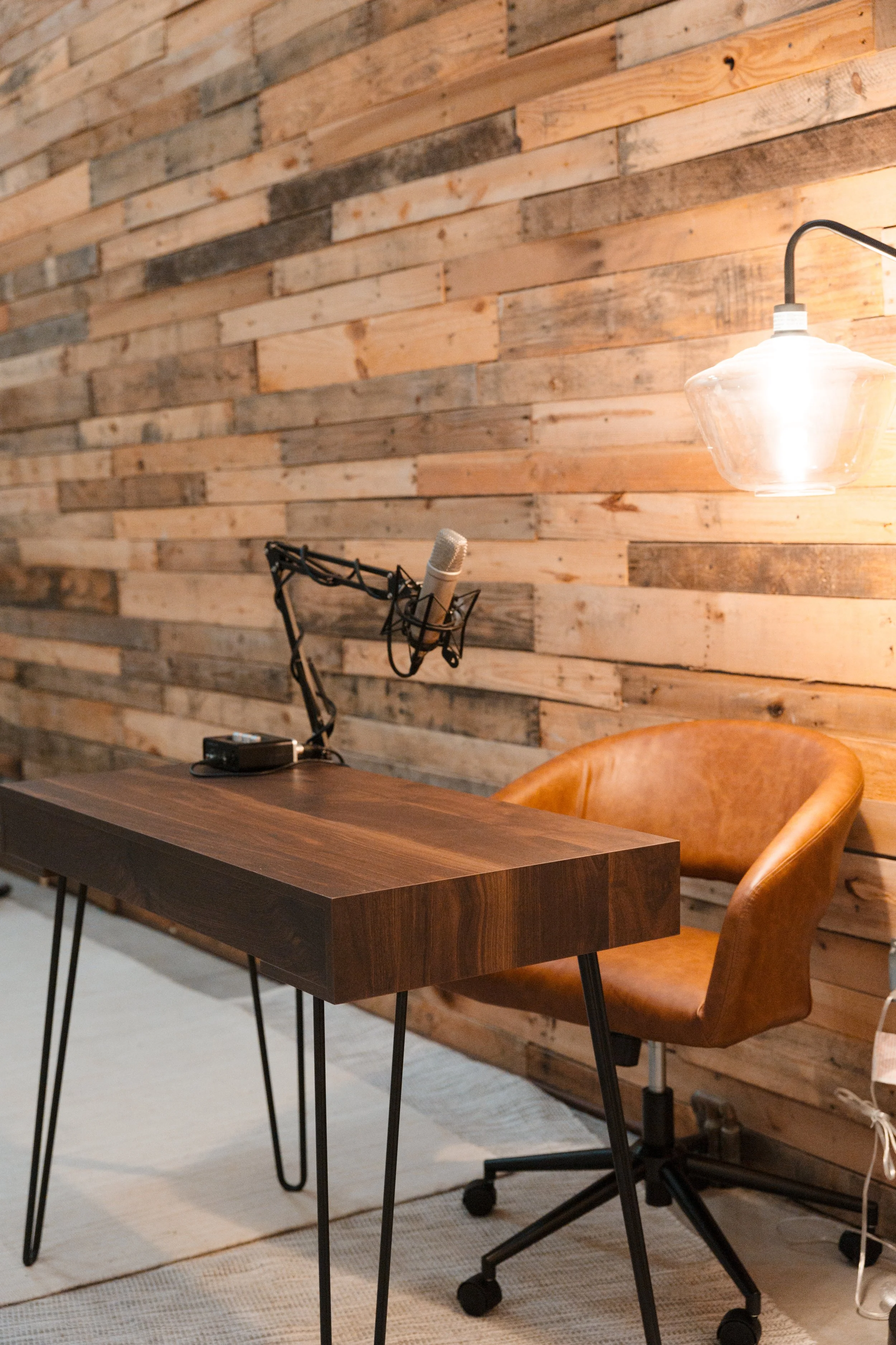 A recording studio or podcasting space with a wooden wall, a brown office chair, a wooden desk, a microphone on an adjustable stand, and a hanging glass light fixture.