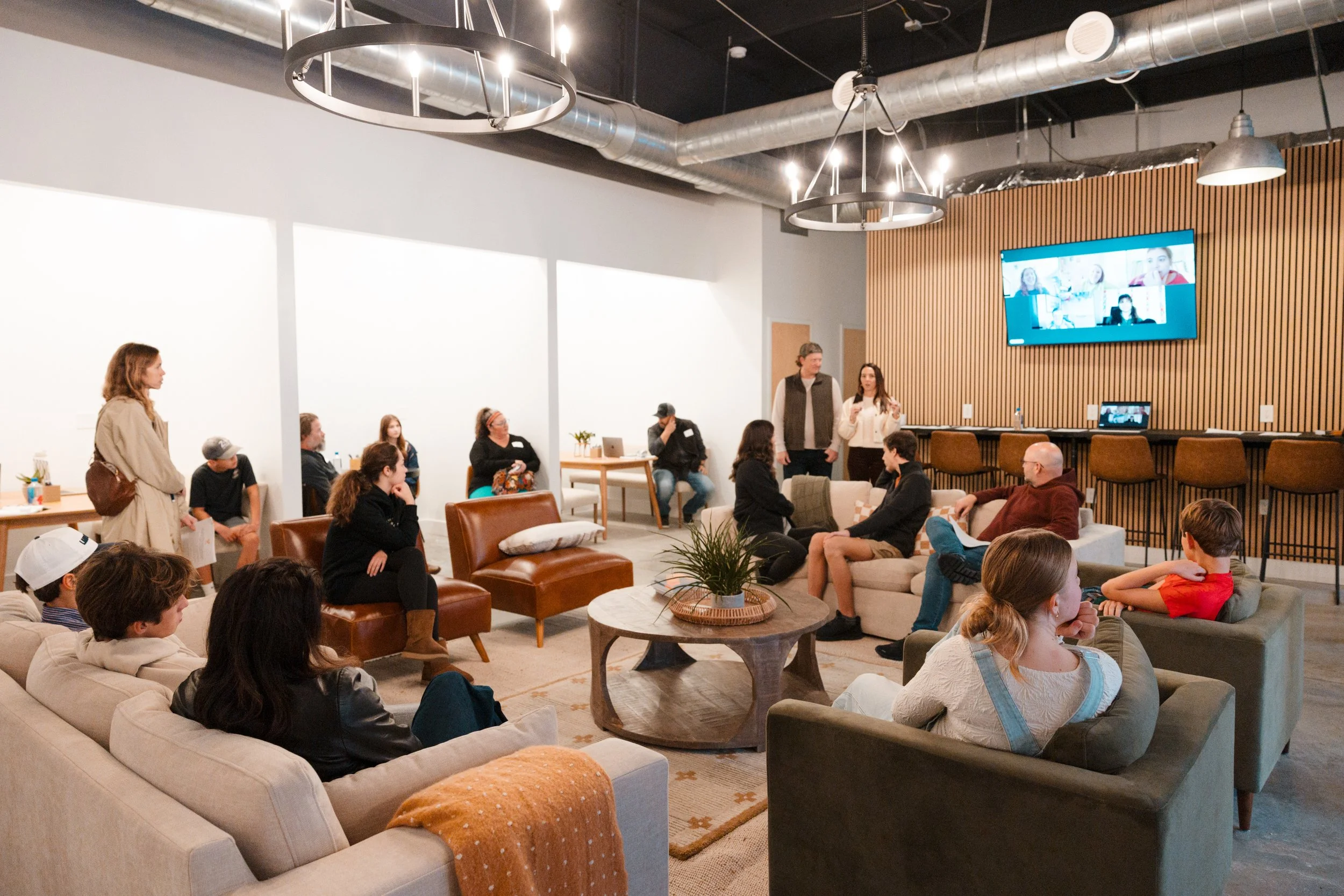 Group of people in a modern lounge or living room attending a presentation or meeting with a large screen on the wall showing remote participants.