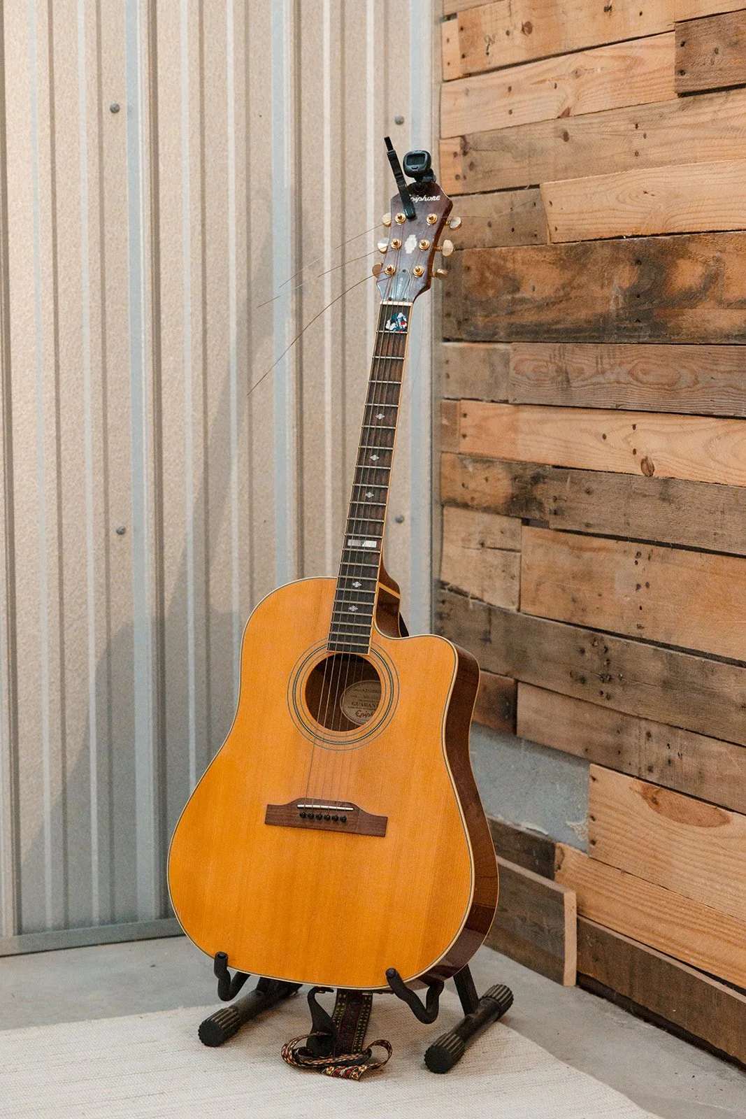 An acoustic guitar on a stand next to a corrugated metal wall and a wooden wall with horizontal planks.