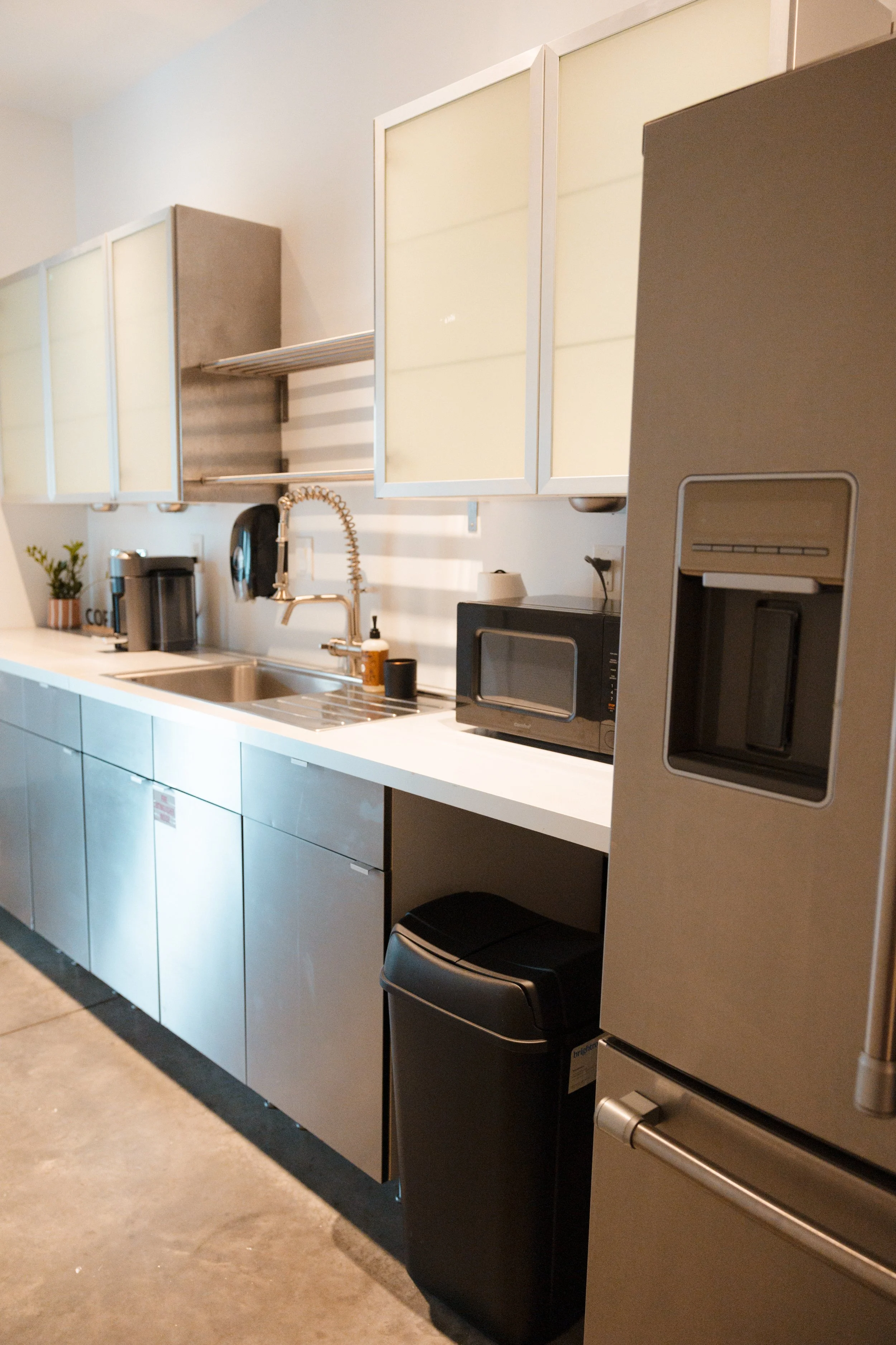 Modern kitchen with a stainless steel refrigerator, a black trash bin, white cabinets, a microwave, a coffee maker, and a sink with a curly faucet.