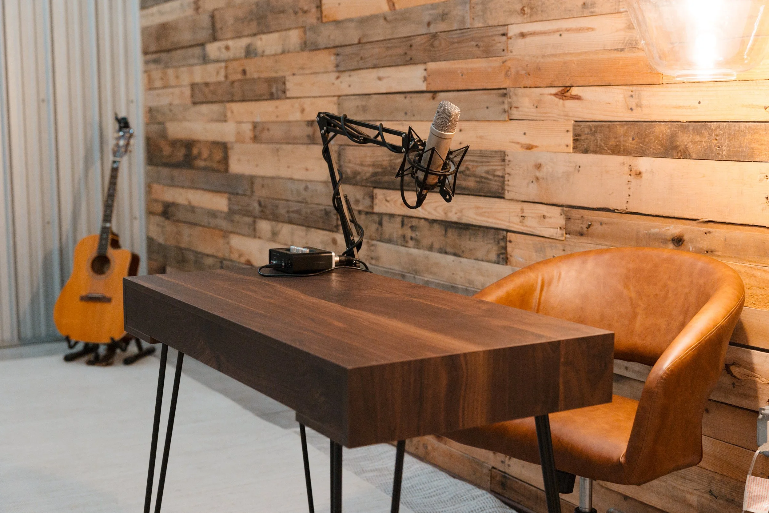 A recording studio with a wooden desk, a professional microphone, a leather chair, and a guitar in the background against a wooden wall. podcasting classes and courses