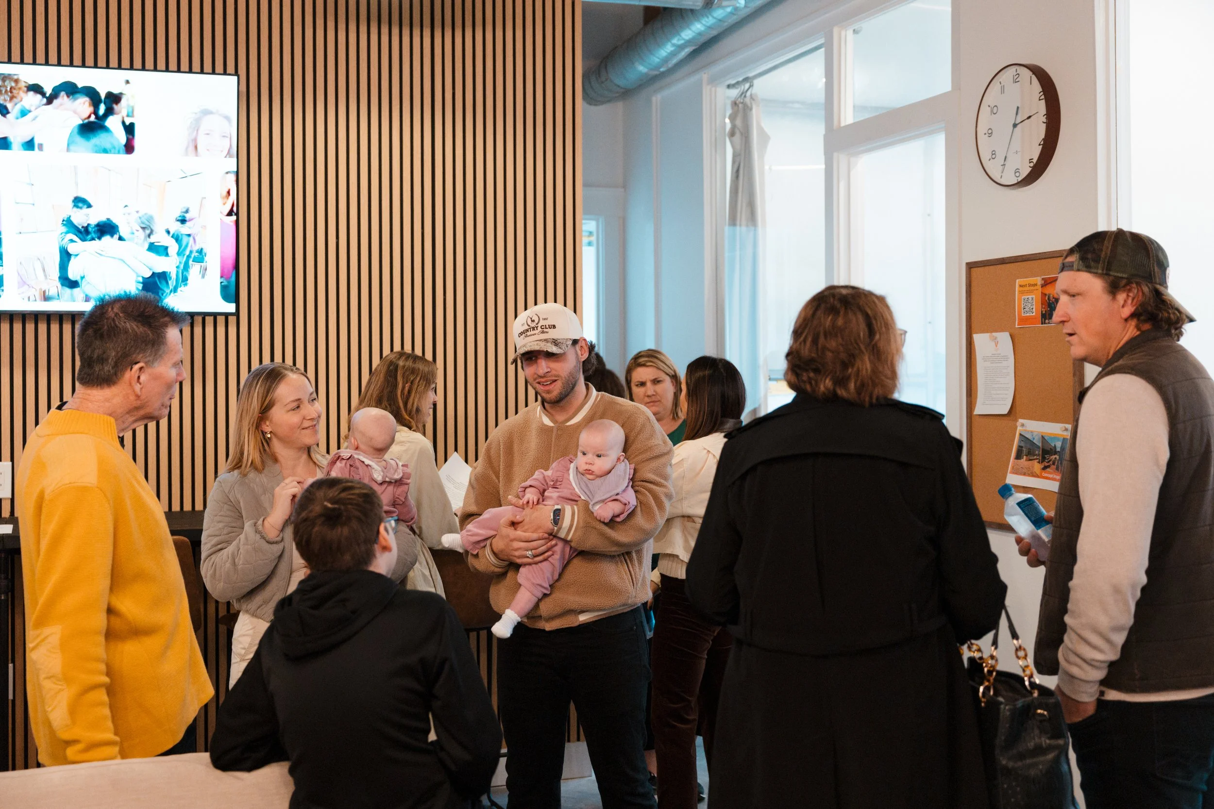 Group of people talking in a lobby, some holding babies, with a large clock on the wall and a bulletin board nearby.