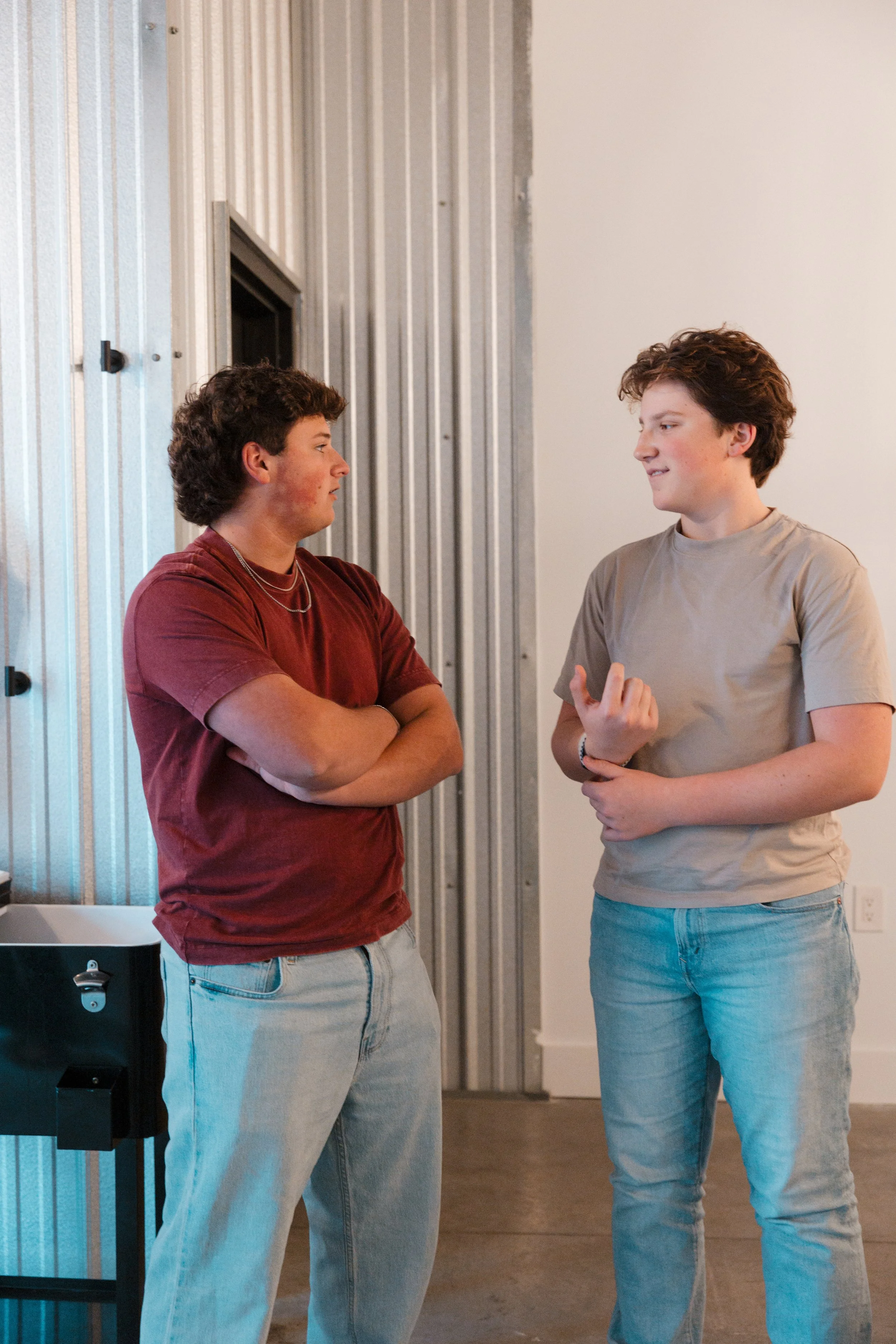 Two young males having a conversation indoors, with one crossing his arms and the other gesturing with his hand, near a wall with metal paneling.
