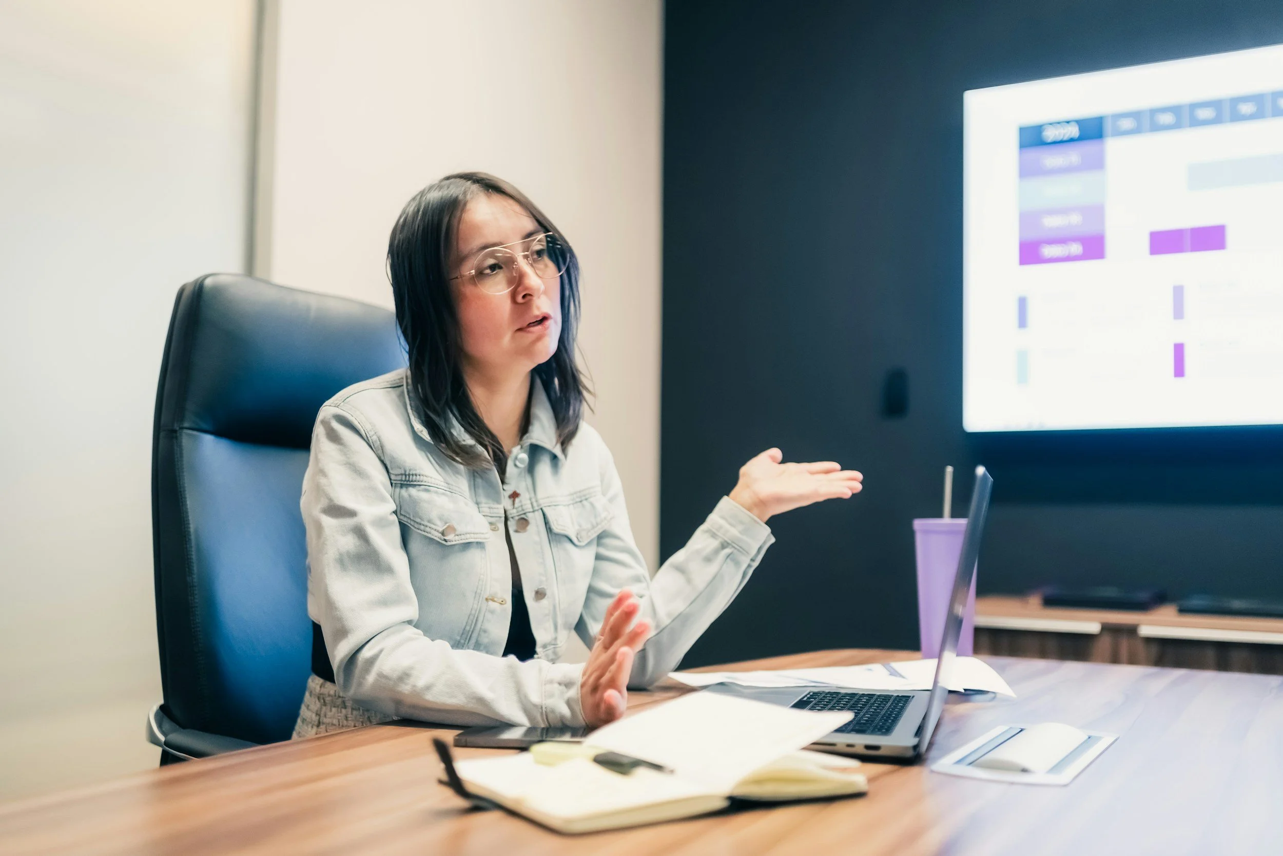 A woman with glasses, wearing a denim jacket, sitting at a desk in a conference room, gesturing with her left hand while looking at a large screen displaying a colorful chart or schedule.