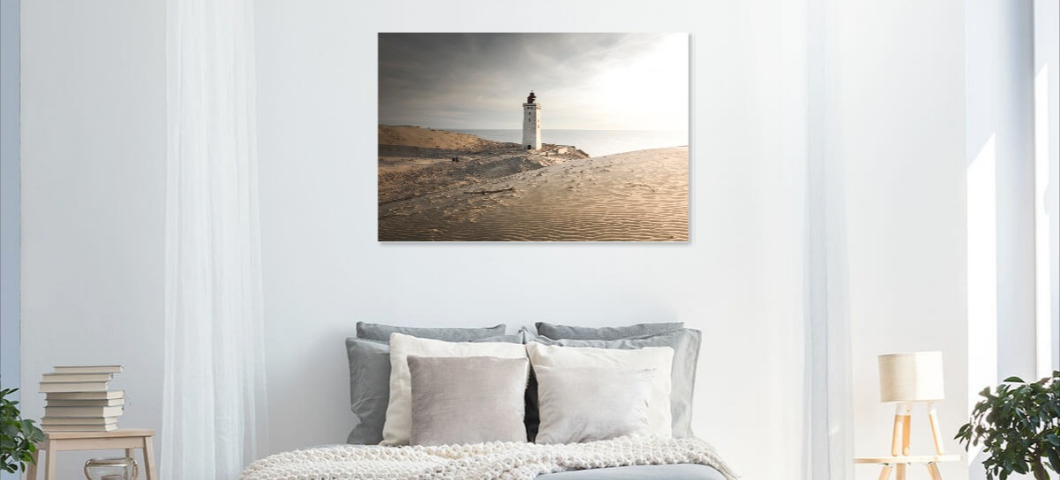 A bedroom with white walls and large windows, featuring a bed with gray and cream pillows, and a painting of a lighthouse on a sandy shore hanging above the bed.