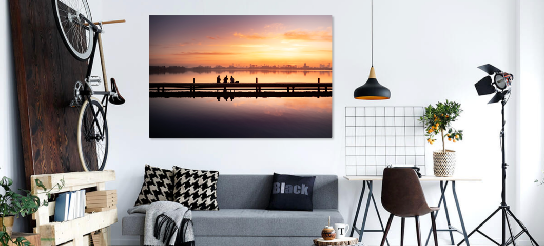 Modern living room with a gray sofa, patterned pillows, and a blanket. Wall art features a sunset over a lake with a dock and people. Bicycle mounted on a wooden wall panel, bookshelf with books, and a potted plant. A desk with a chair, a grid displa