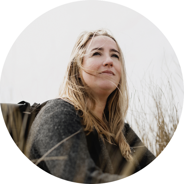 A woman with long blonde hair, wearing a dark gray sweater, sitting outdoors among tall grass, looking up with a contemplative expression.